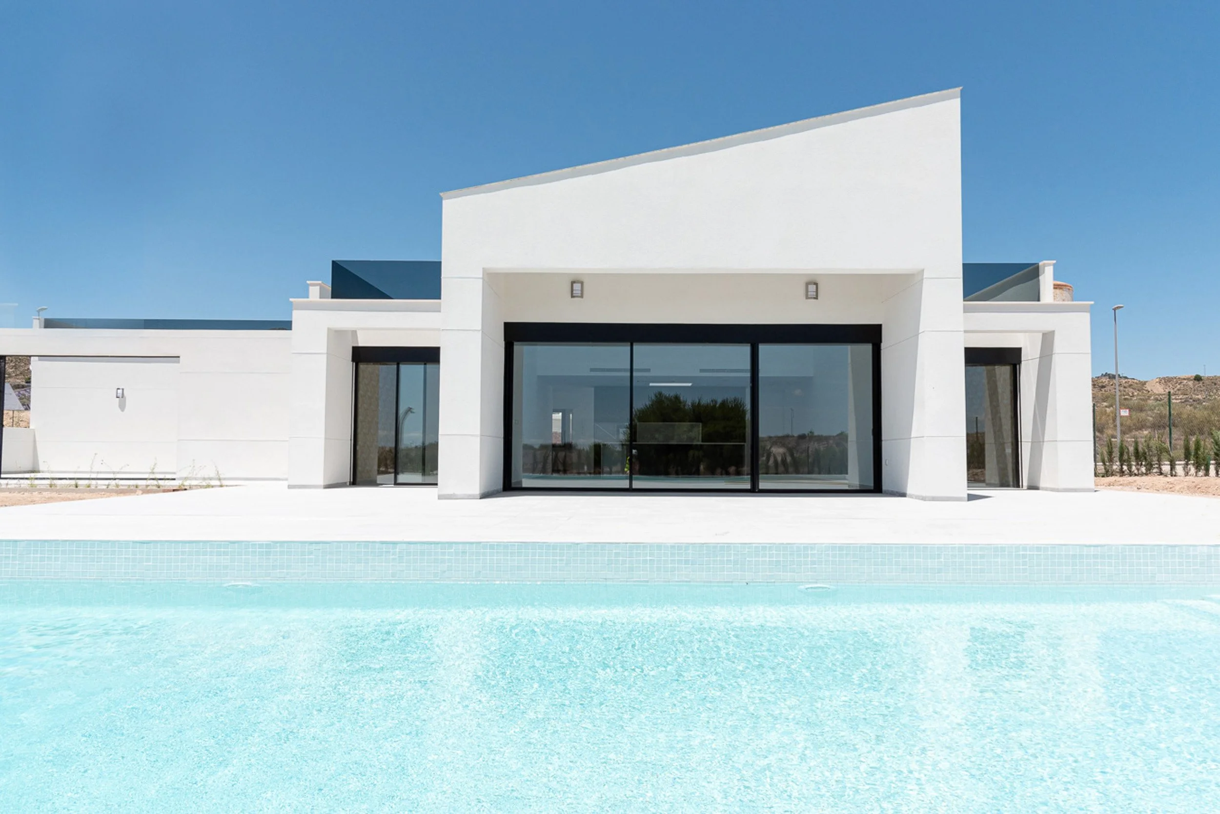 Casa moderna de color blanco con grandes ventanas y puertas de cristal, en un ambiente soleado con un piscina en primer plano y un cielo despejado.