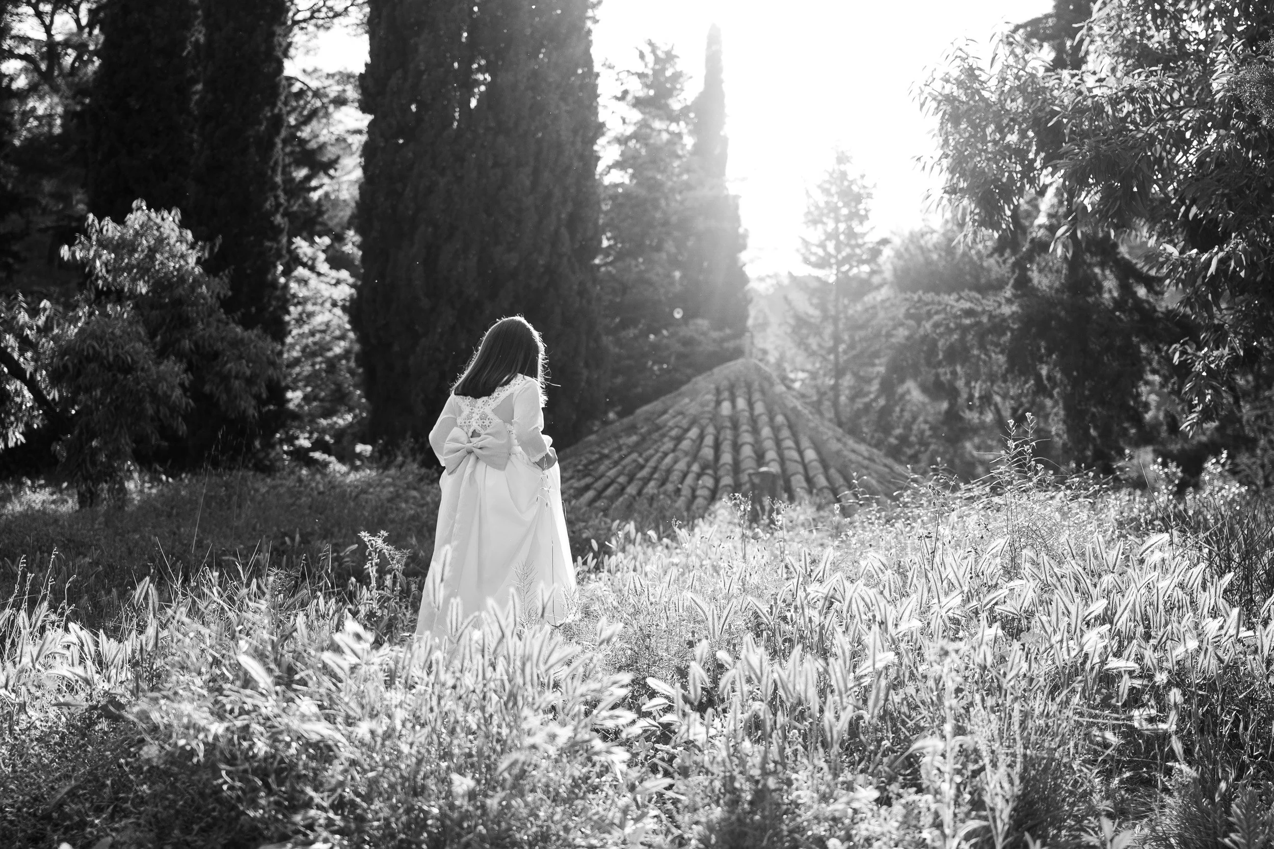 Niña con vestido de comunión andando en un campo, de espaldas, rodeada de árboles, en blanco y negro.