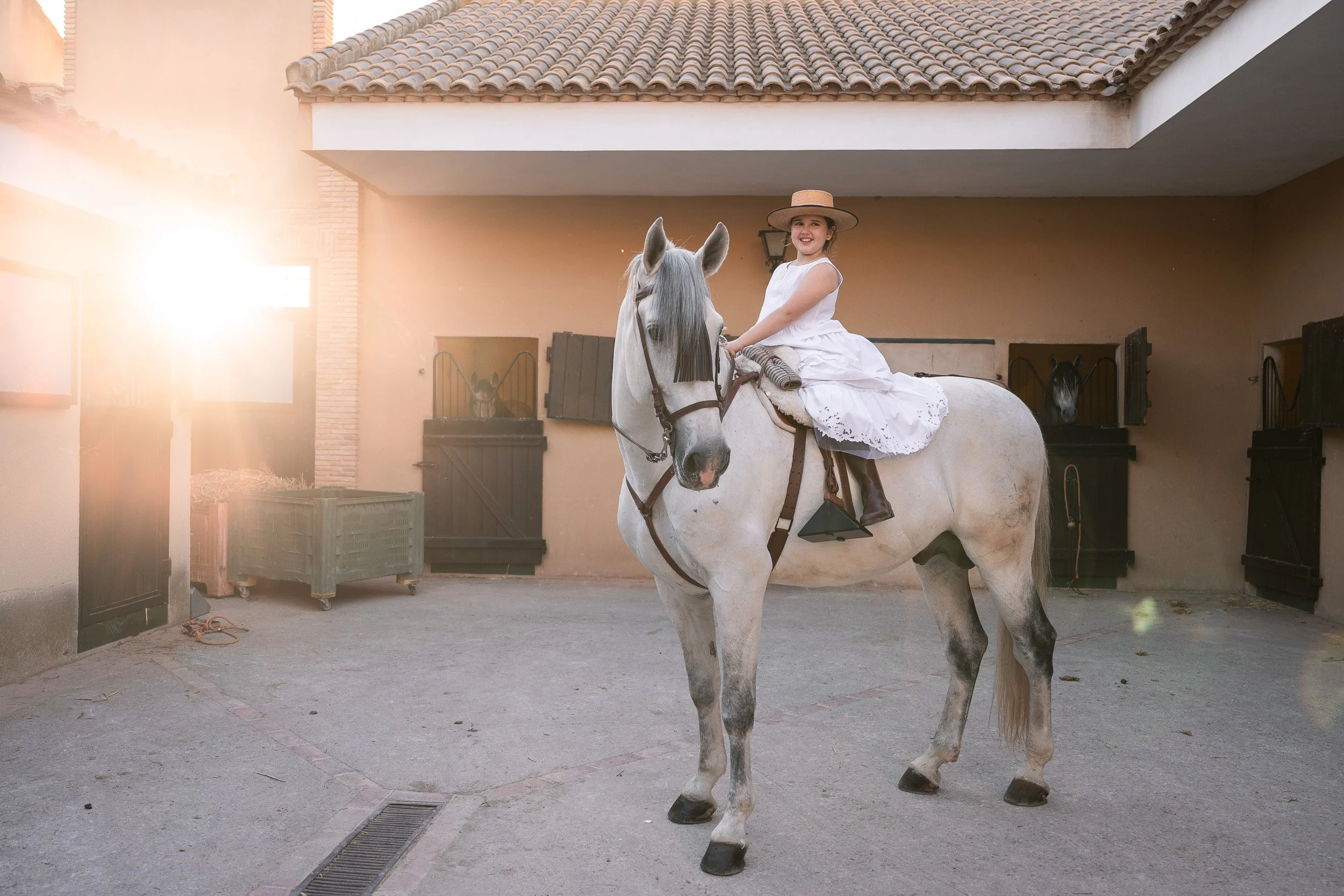 Una niña vestida con un vestido blanco y un sombrero grande monta un caballo gris en un establo bajo luz solar y con un fondo de paredes en color terracota y cajas de transporte.