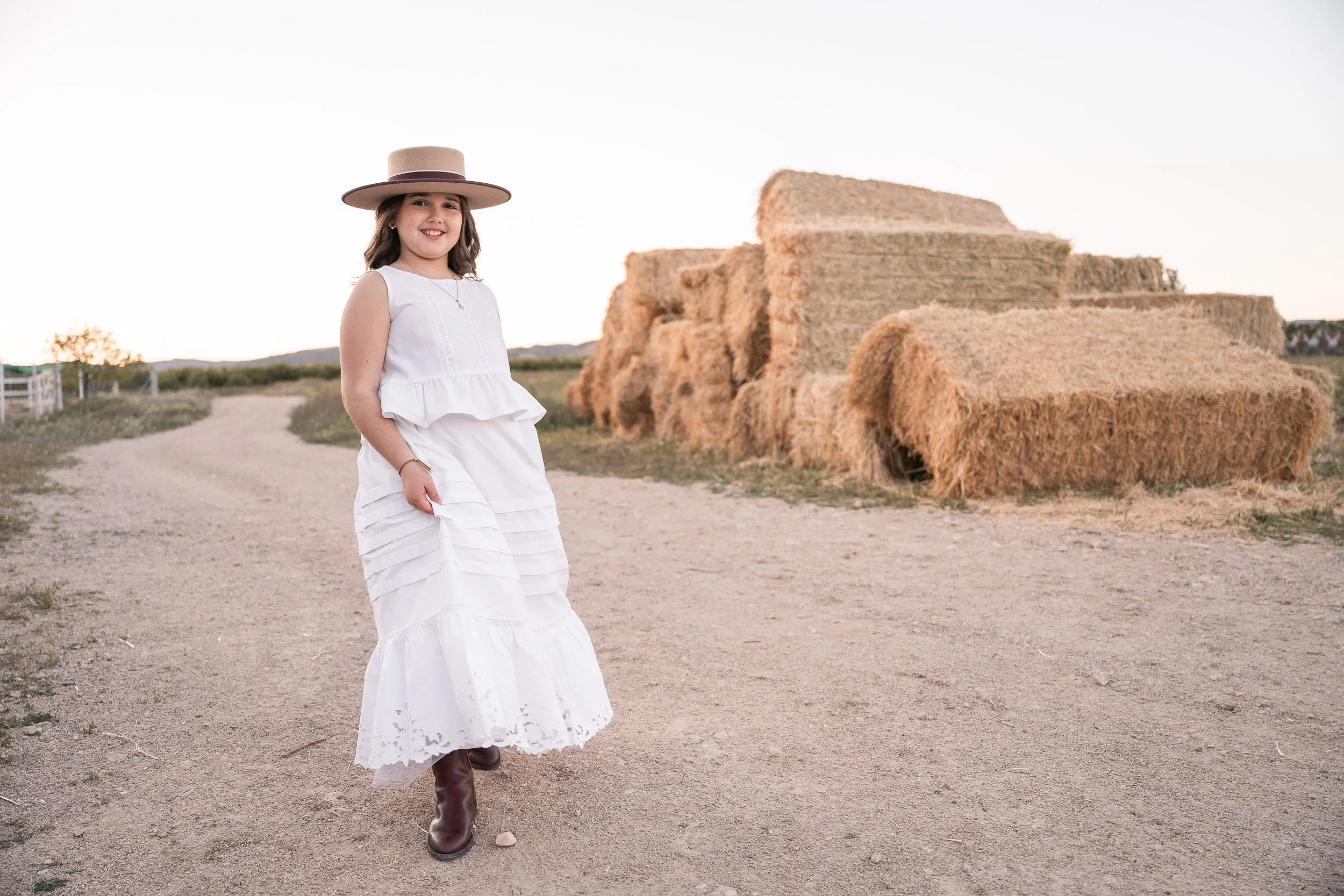 Joven niña con vestido blanco y sombrero grande caminando por un camino de tierra en un campo con pacas de heno en el fondo al atardecer.