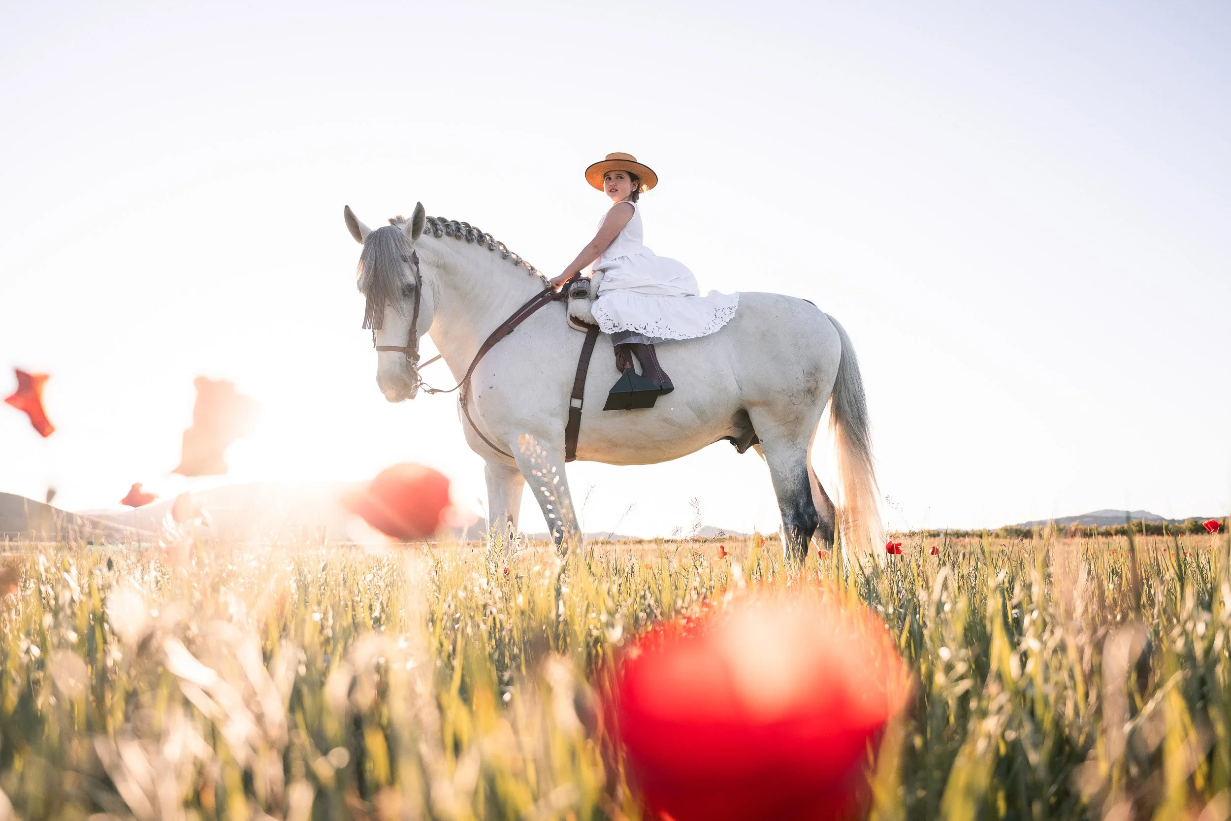 Niña de comunión montada en un caballo en un campo con flores y un cielo despejado al atardecer.