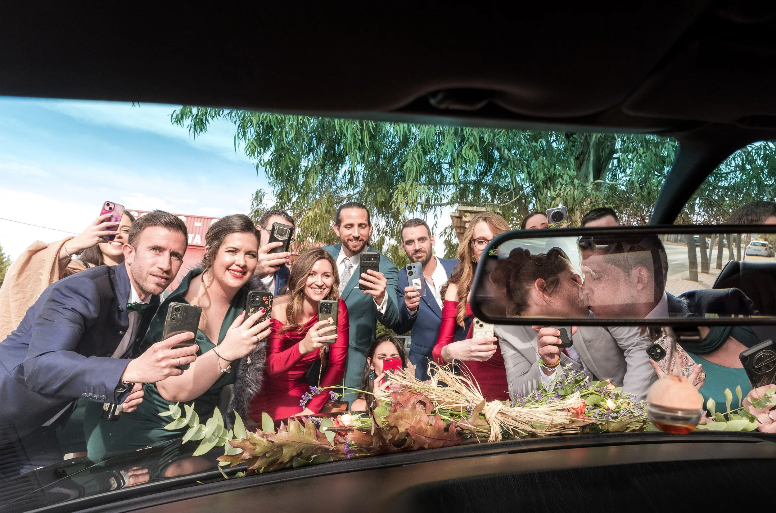 Grupo de personas de aspecto feliz y diverso tomando fotos y sonriendo desde un vehículo, con decoración de hojas y flores en el interior.