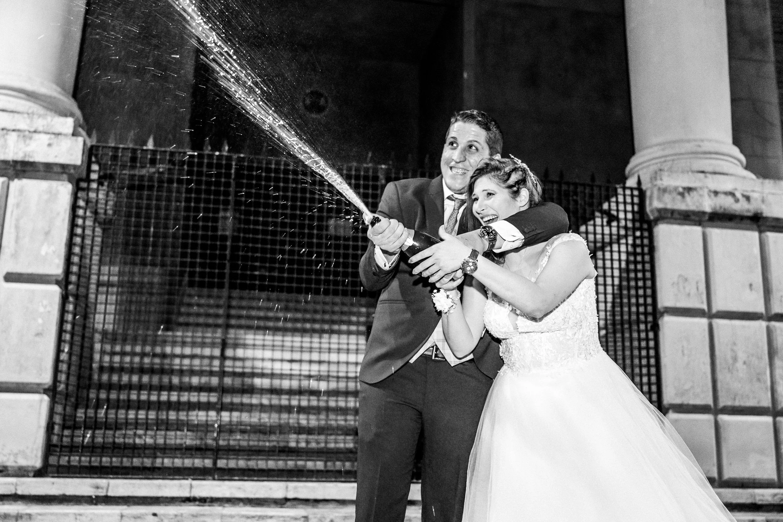 Pareja de novios en su boda brindando con champagne en un escenario al aire libre, con columnas y una cerca de fondo.