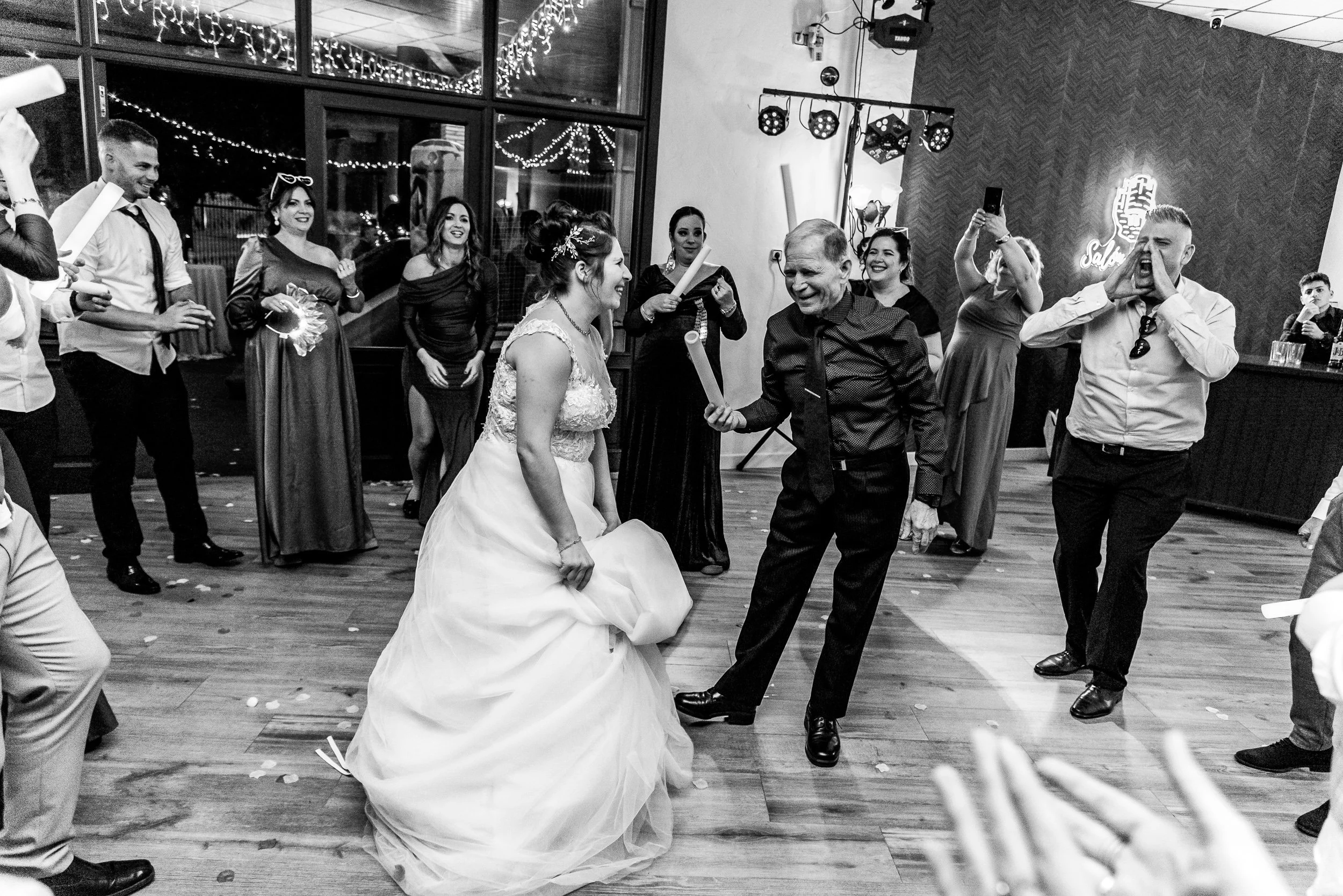 Pareja de recién casados bailando en una recepción. Rodeados de amigos y familiares, con decoraciones festivas y luces de fiesta en el fondo.