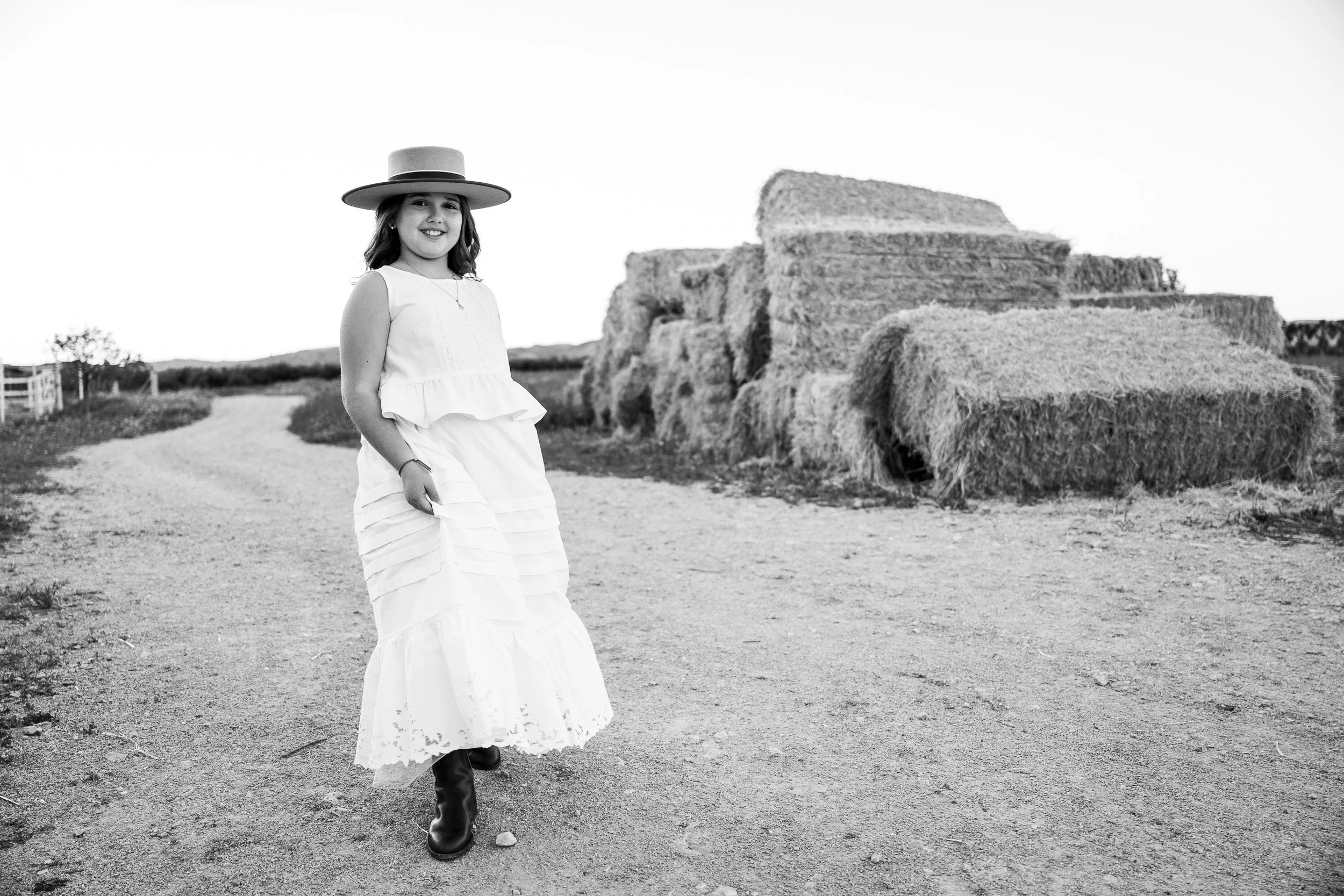 Una niña con enaguas, sombrero y botas negras caminando por un camino en un campo, con pilas de paja, en un día soleado y despejado.
