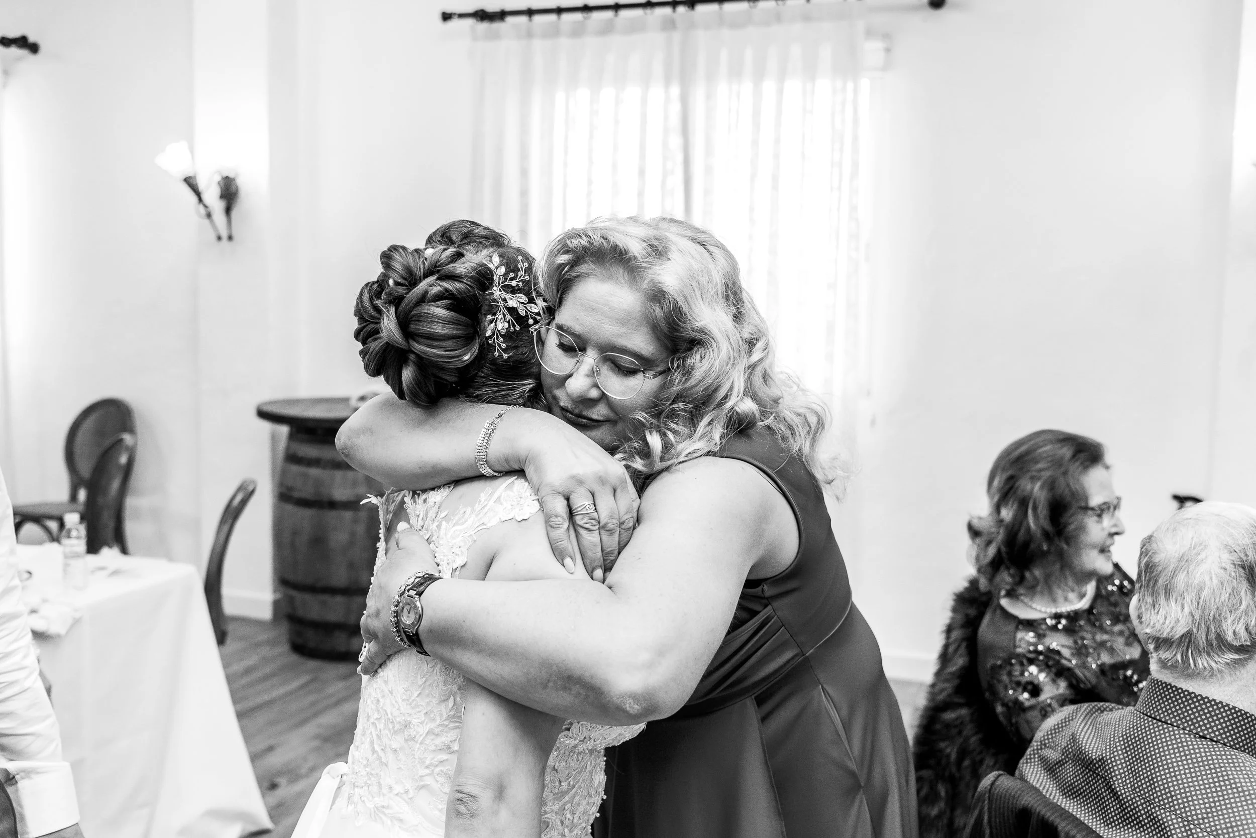 Dos mujeres abrazándose en celebración, una de ellas lleva un vestido de novia y la otra un vestido sin hombros, en un ambiente de fiesta con personas sentadas y decoraciones.