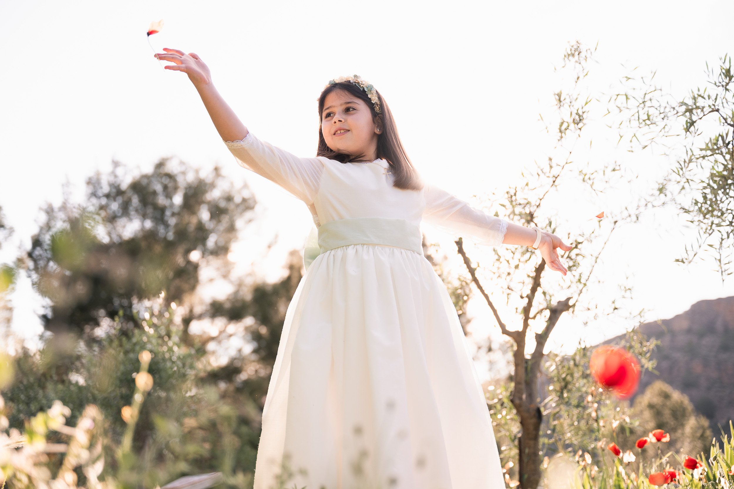 Una niña con vestido de comunión en un campo durante el atardecer, lanzando una flor y rodeada de flores y árboles.