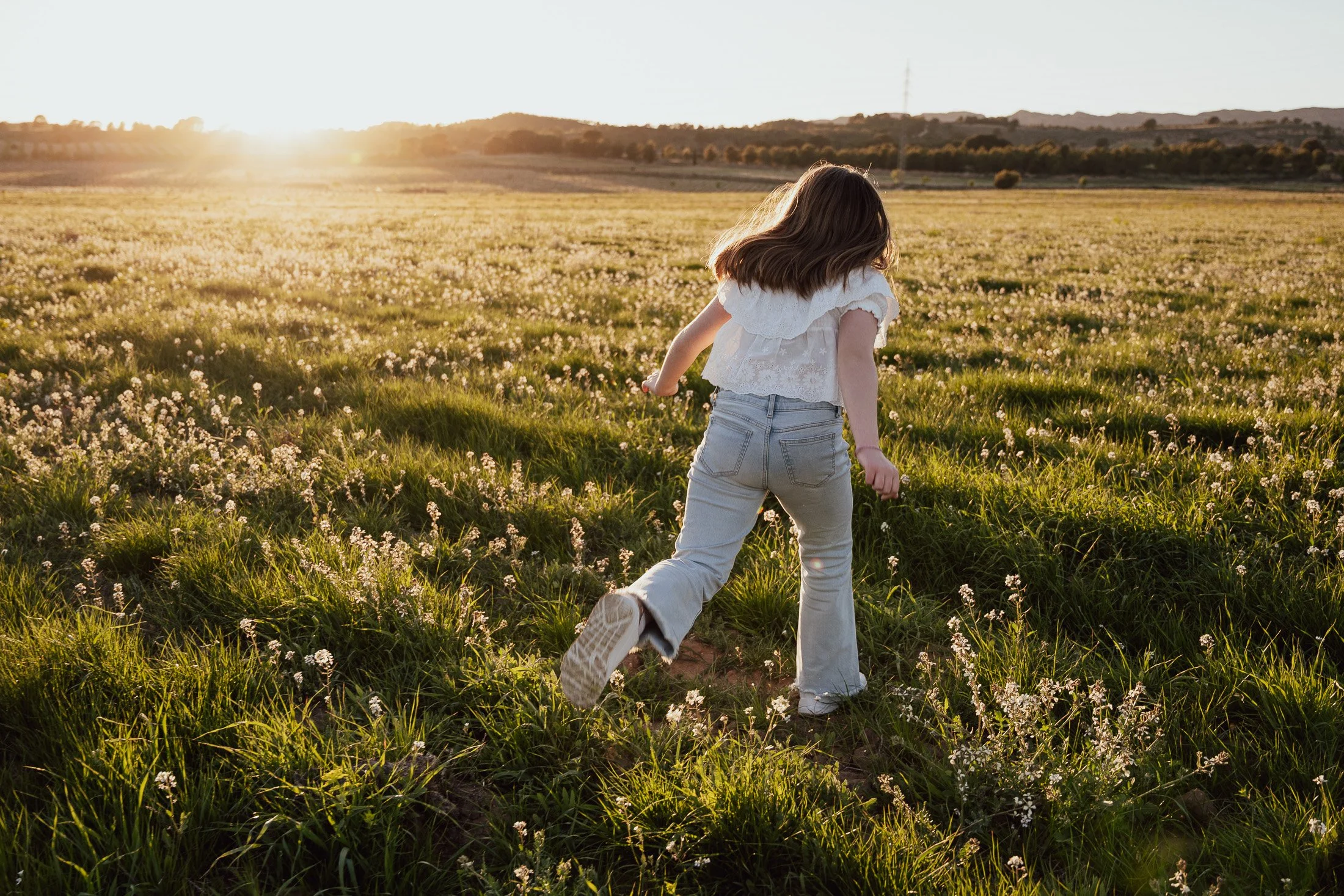 Una niña corriendo por un campo de flores al atardecer, con un paisaje de colinas y árboles en el fondo.