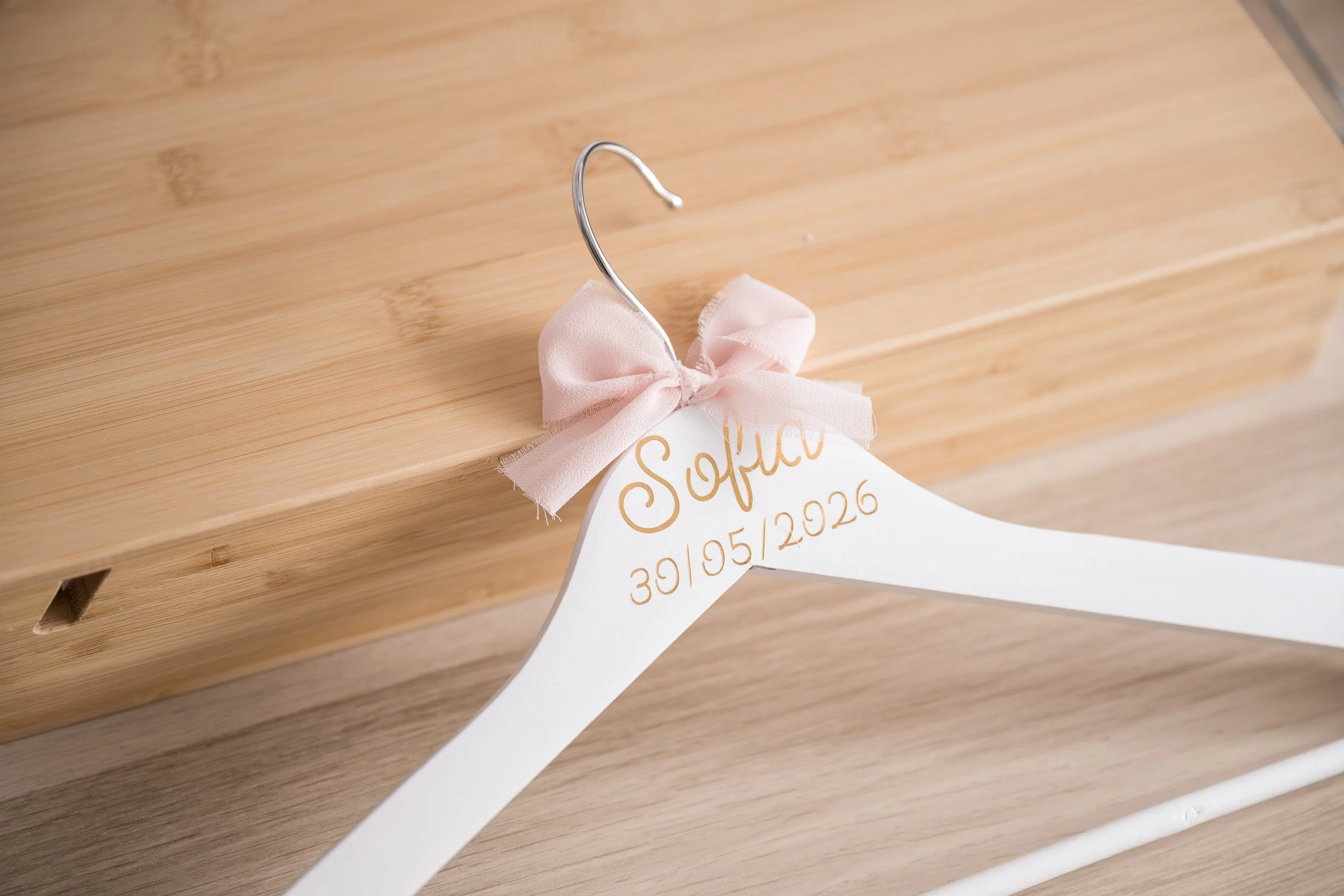 Gancho decorativo de metal con lazo de tela rosa, sujetando una tarjeta blanca con detalles dorados y texto en español, colgado de una superficie de madera clara.