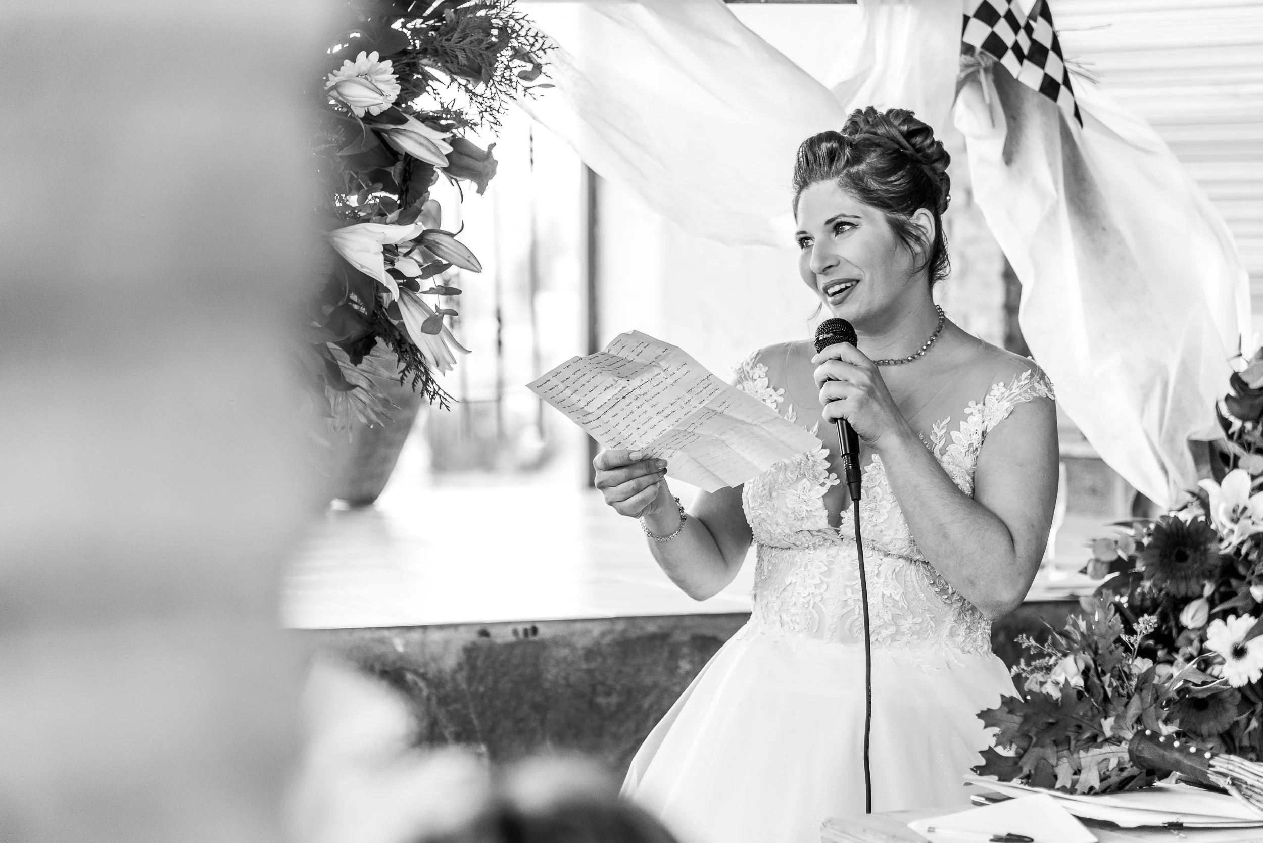 Mujer vestida de novia leyendo en un micrófono en una ceremonia, rodeada de flores.