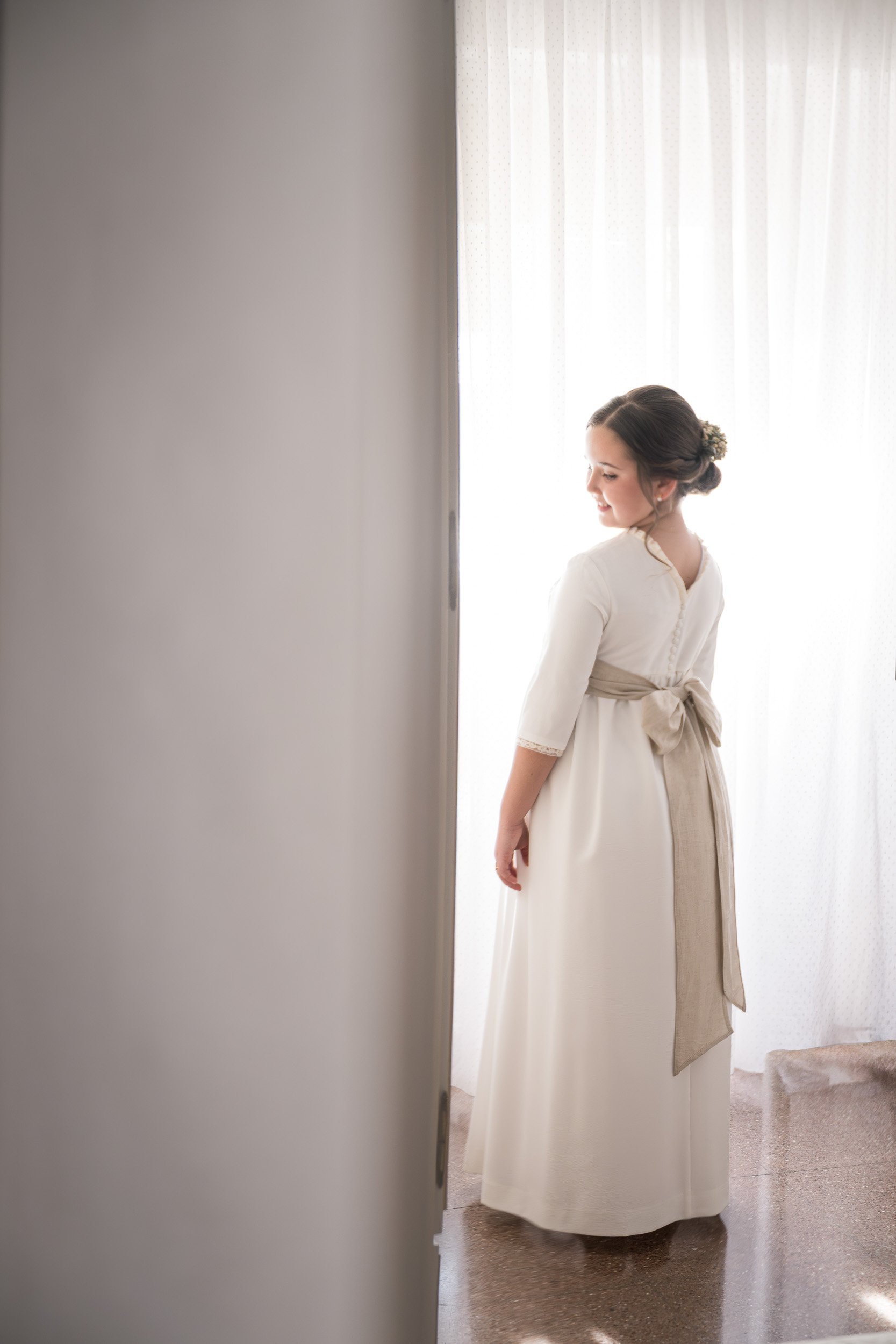 Mujer vestida con un vestido largo y de color crema, con cabello recogido adornado con flores, de espaldas, en un entorno con cortinas blancas que filtran la luz natural.