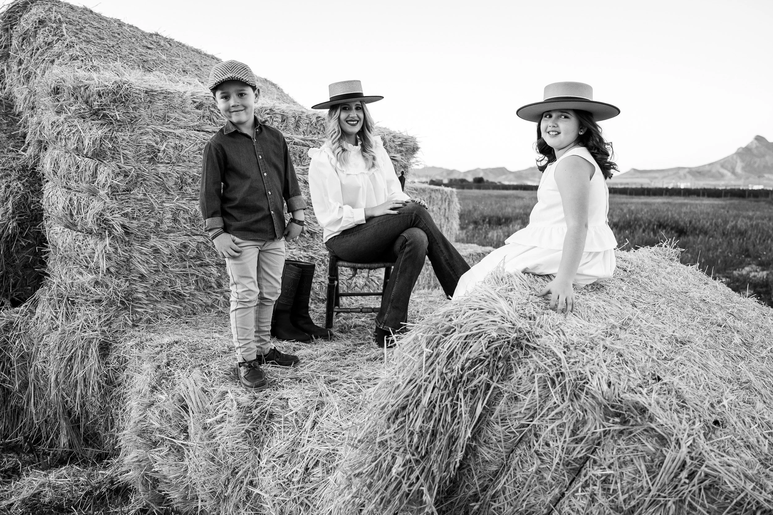 Tres personas, dos mujeres y un niño, en un campo con pacas de heno, todos sonriendo y usando sombreros grandes en un ambiente rural