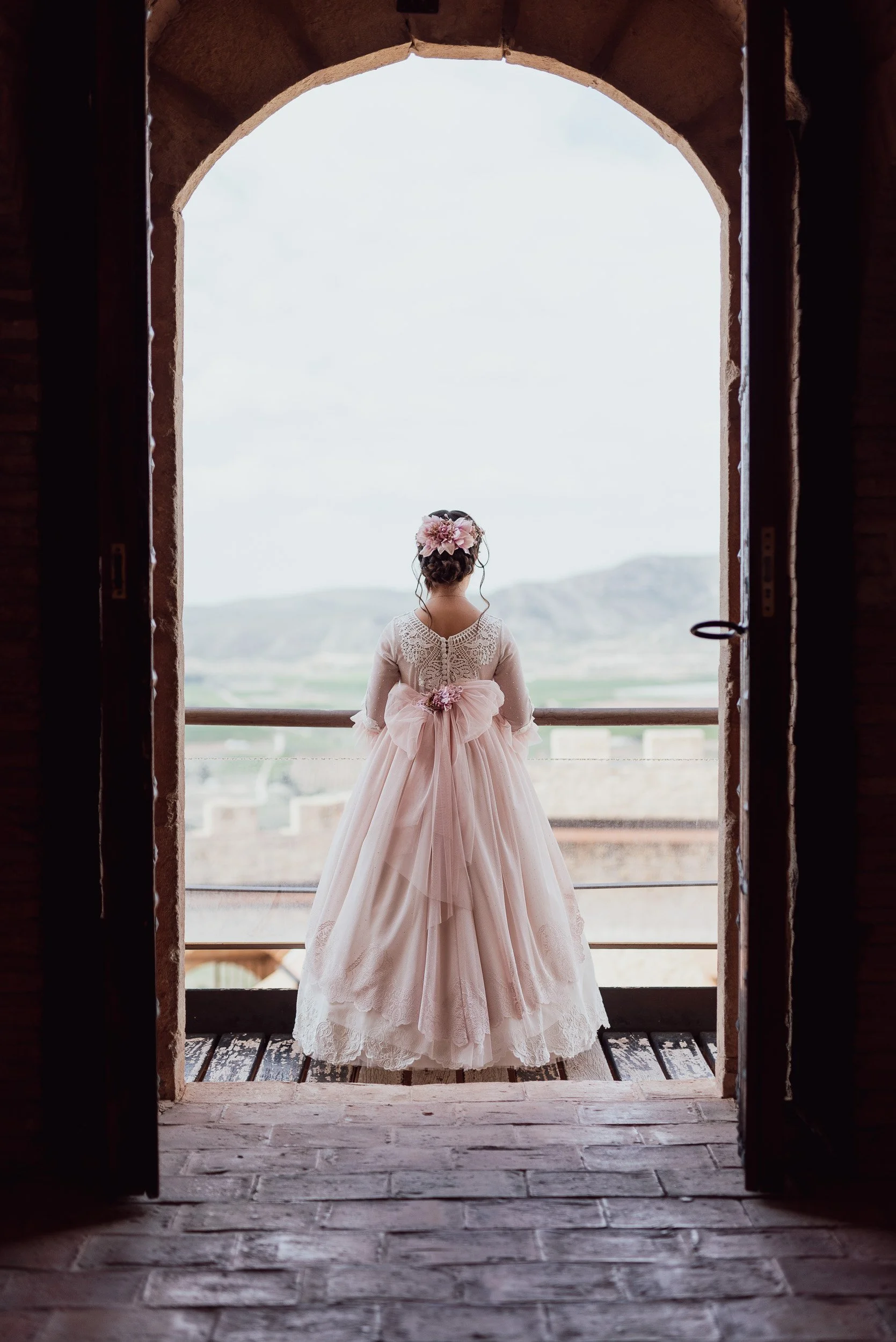 Niña con vestido de comunión de espaldas, mirando hacia el horizonte en una puerta del castillo.