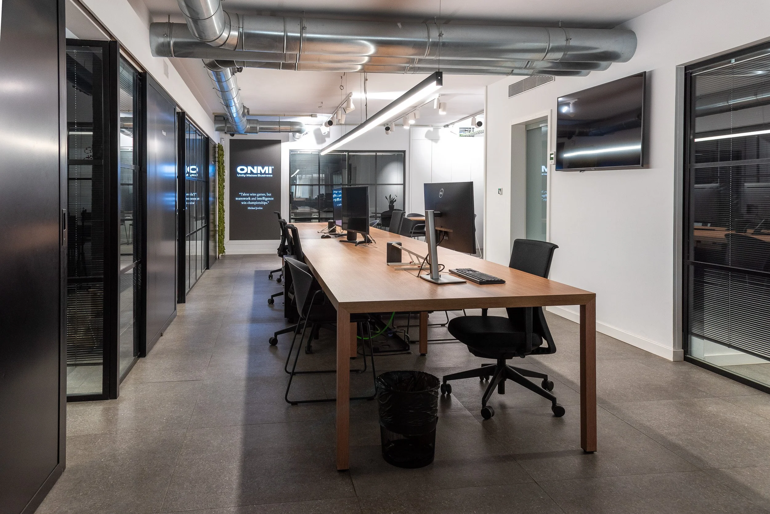 Una sala de oficina moderna con mesas de trabajo, varias computadoras, sillas y paredes de vidrio con cortinas, techo con ductos metálicos expuestos y luces. Pantallas de televisión y un cartel con el logo ONMI.