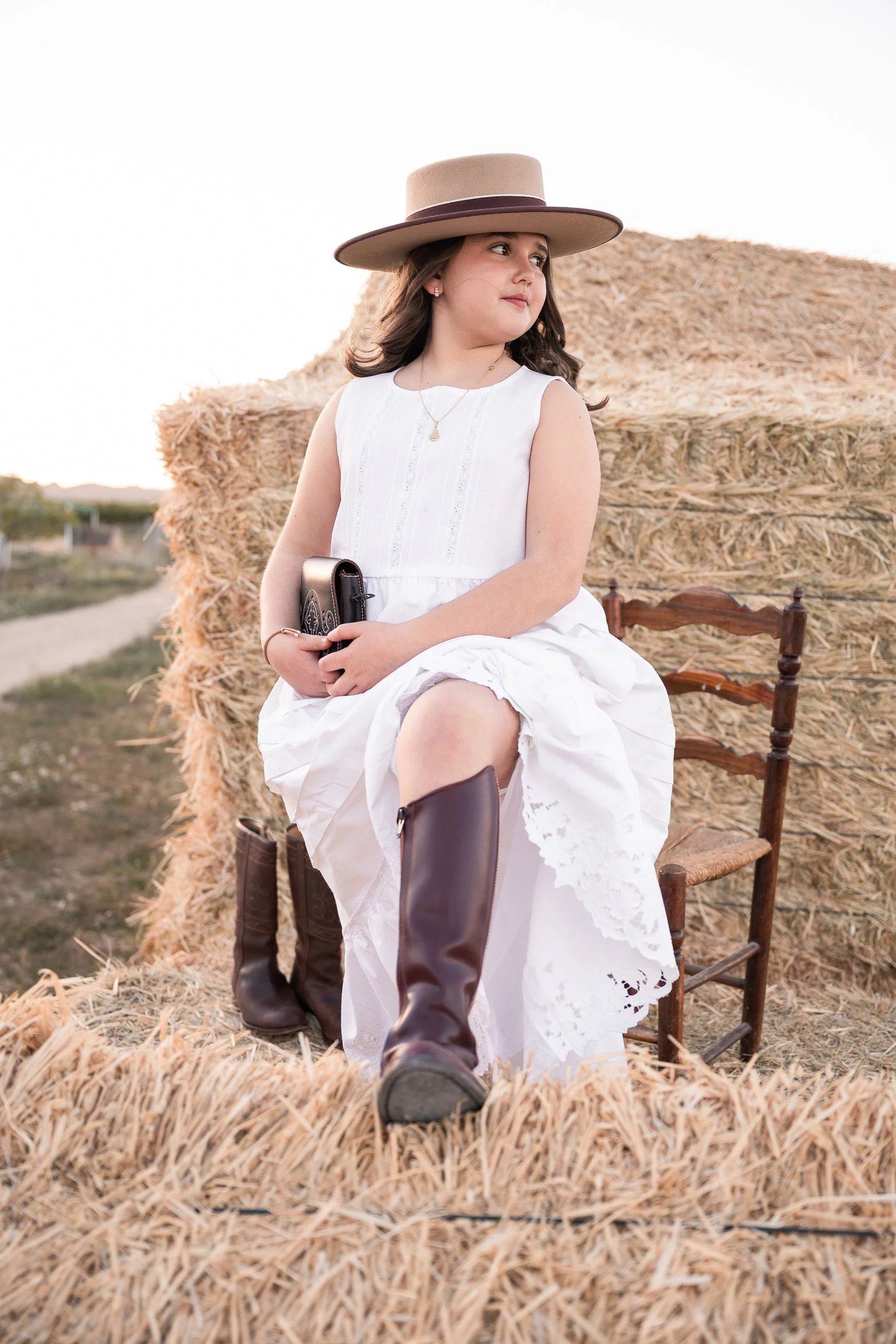 Niña adolescente sentada en un banco de madera entre paja, vistiendo vestido blanco, sombrero ancho y botas de montar, con un paisaje rural de fondo.