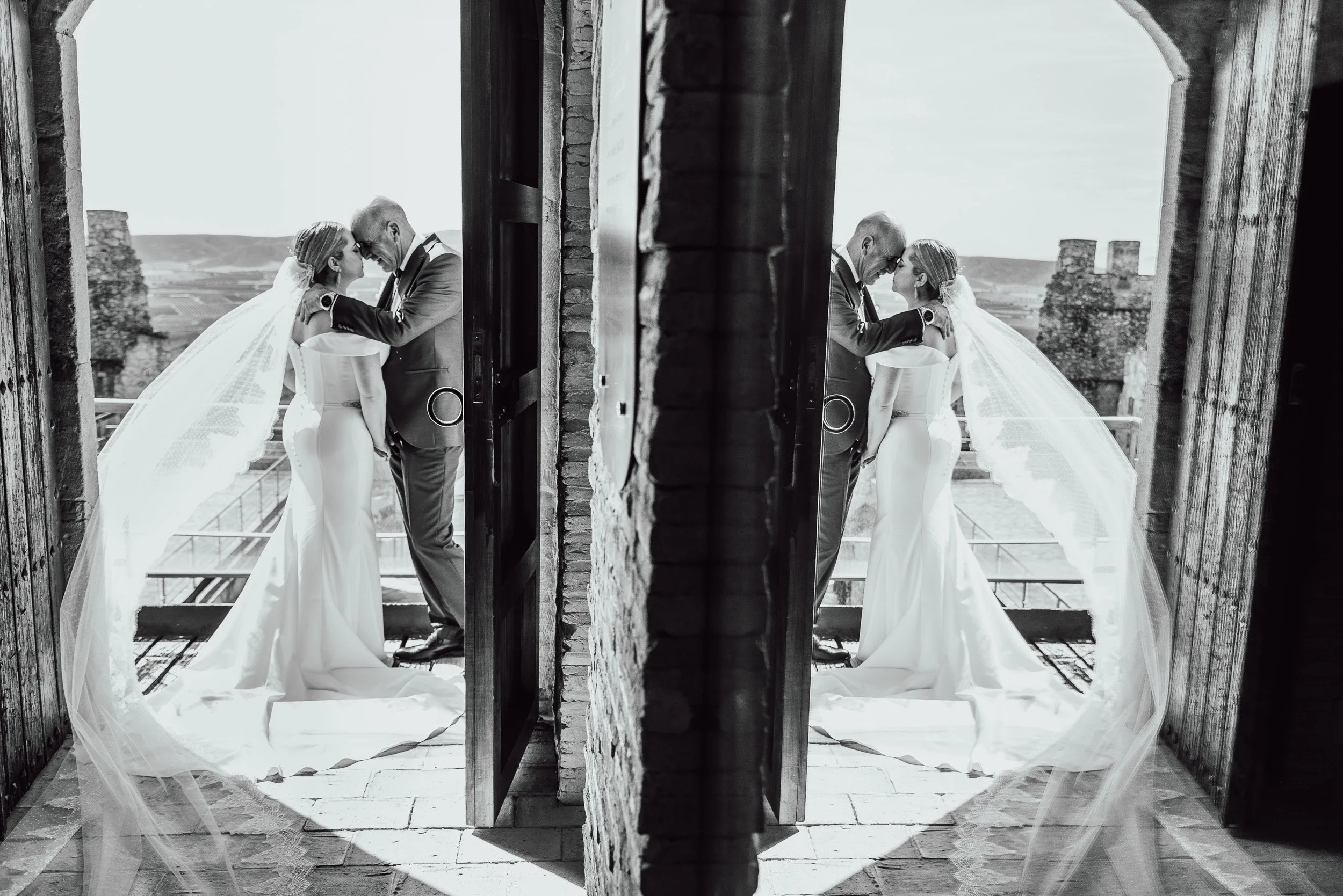 Matrimonio con traje de novios abrazados, reflejados en un espejo, en un entorno al aire libre con paisaje rural al fondo.