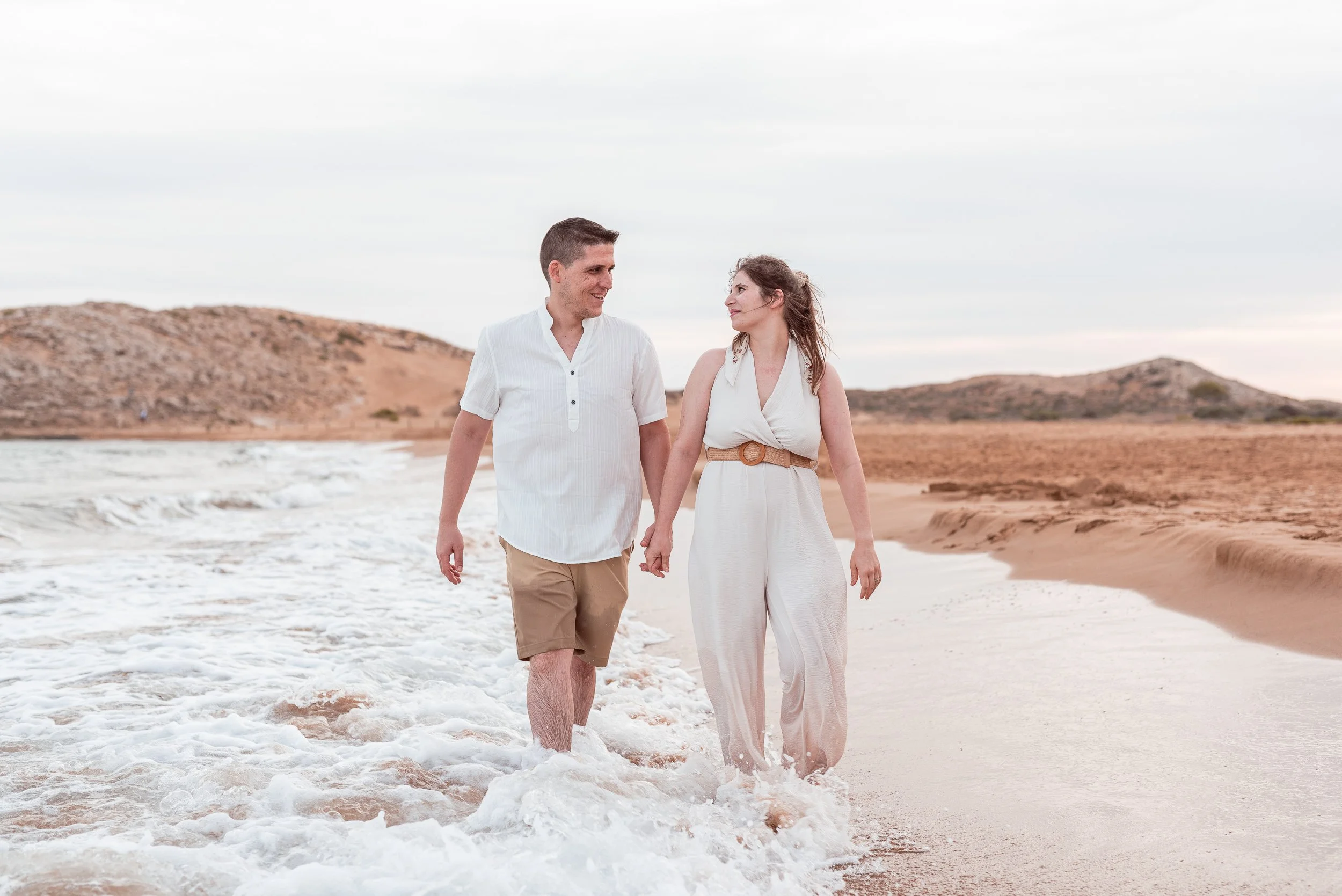 Una pareja caminando por la orilla del mar en una playa, sosteniéndose de la mano y disfrutando del momento. El cielo está despejado y el paisaje es árido con colinas en el fondo.