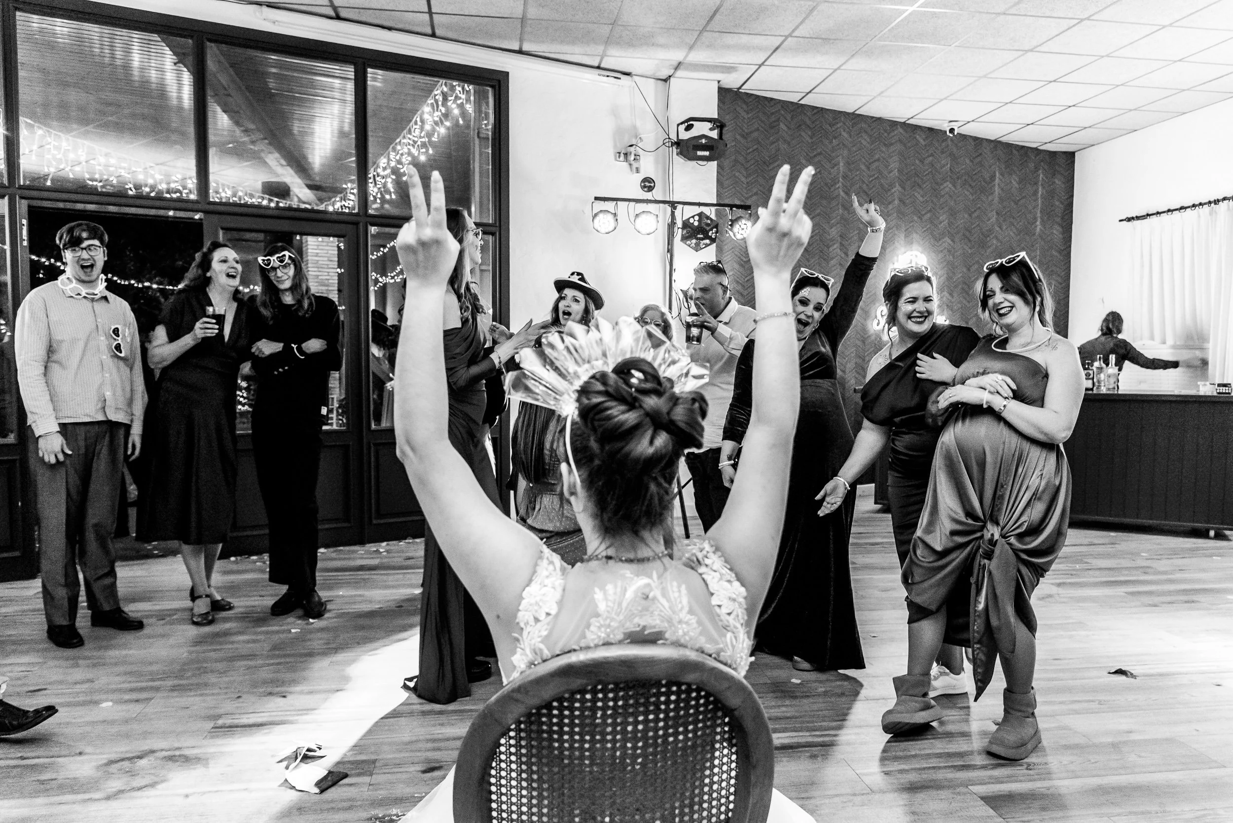 Grupo de personas bailando y divirtiéndose en una celebración o fiesta en un salón, algunas con accesorios como gafas y sombreros, mientras una mujer en primer plano con flores en la cabeza y vestido de encaje baila frente a todos.