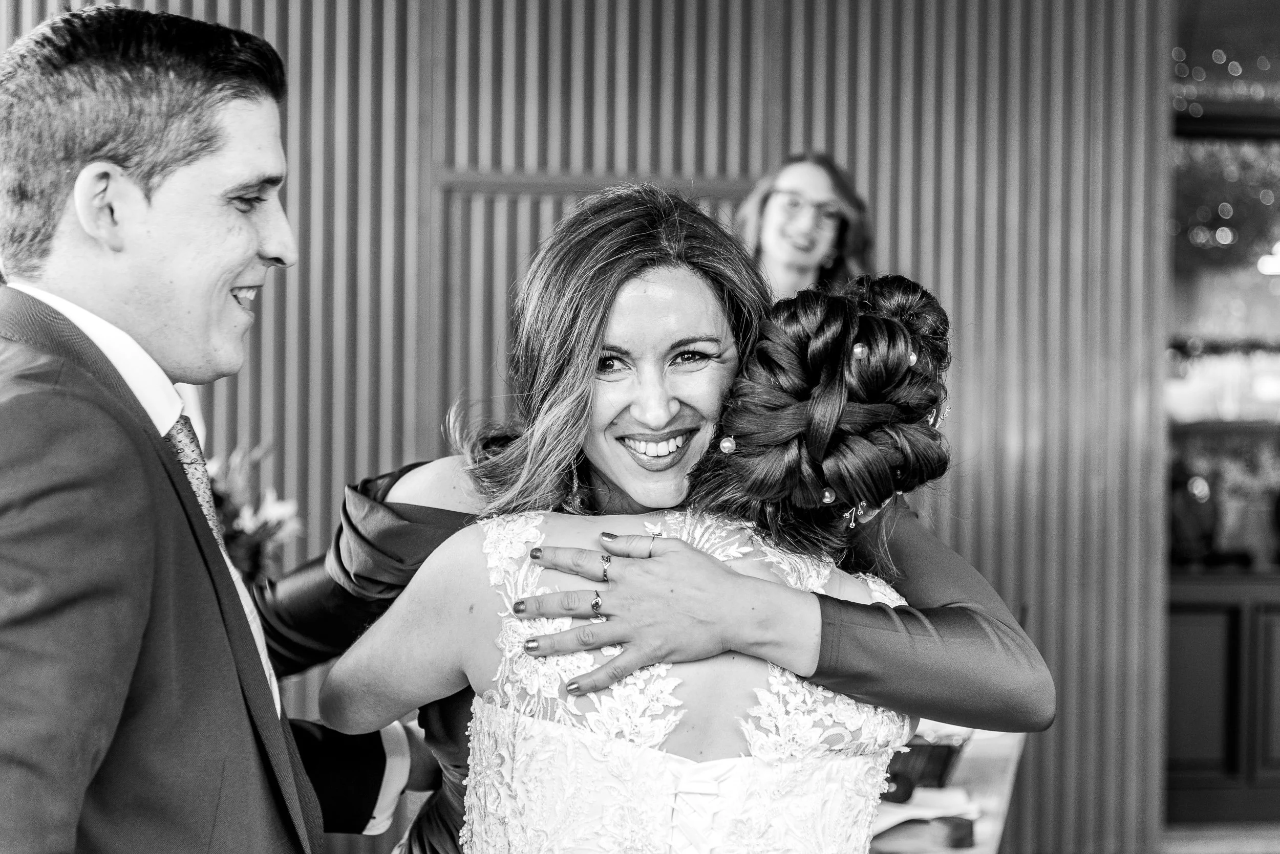 Mujer sonriendo abrazando a otra persona, mientras un hombre le sonríe en un evento, imagen en blanco y negro.