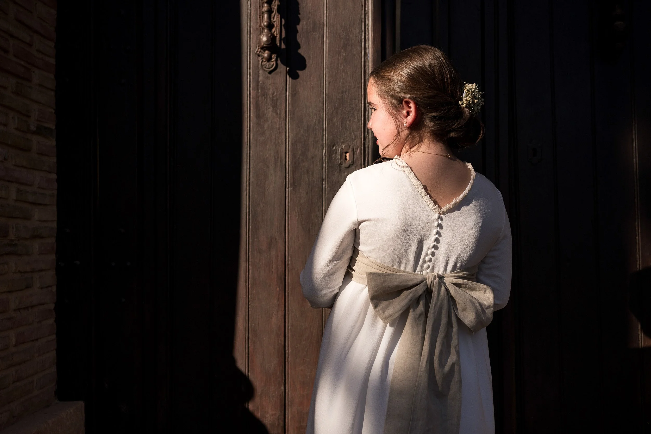 Joven mujer de perfil con vestido blanco, peinado con flores blancas, de espaldas, frente a una puerta de madera y ladrillo, con sombra en el lado izquierdo.