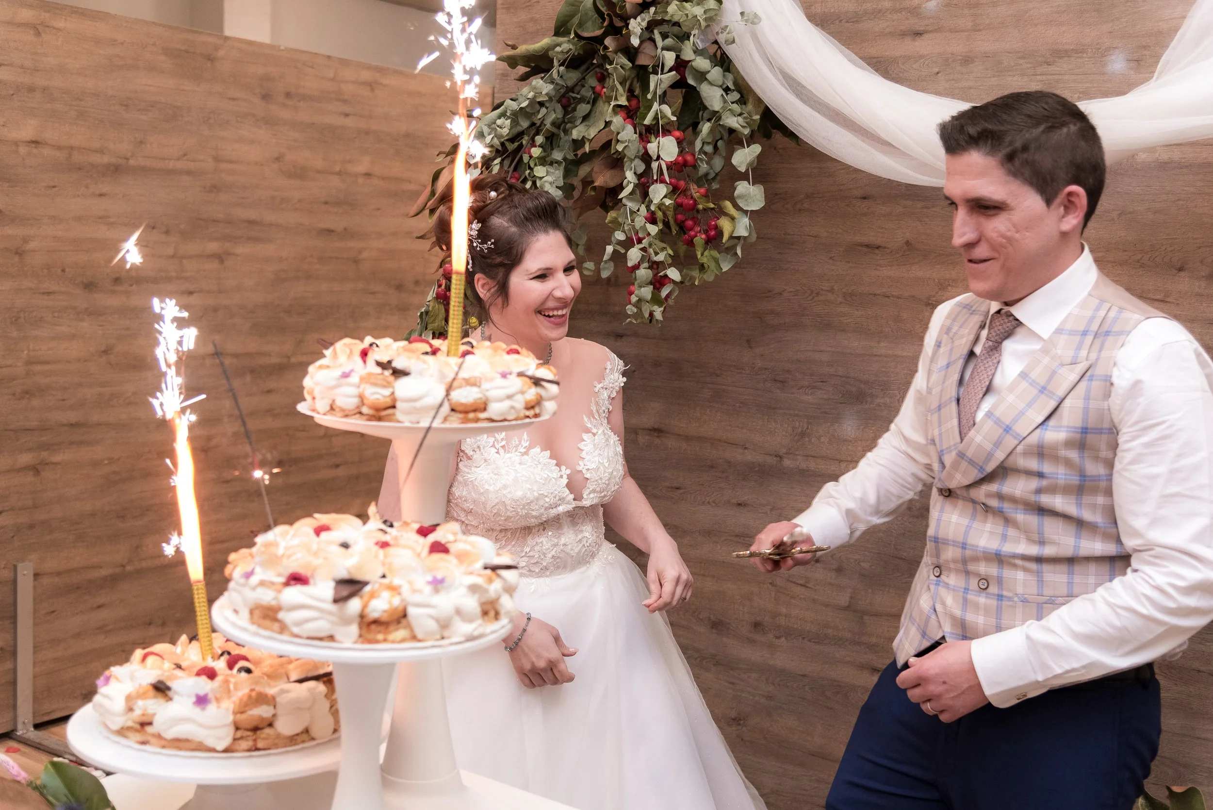 Pareja en vestido de boda, sonrientes, cortando un pastel con dulces, en una celebración de matrimonio. Decoración floral y tela blanca en el fondo.