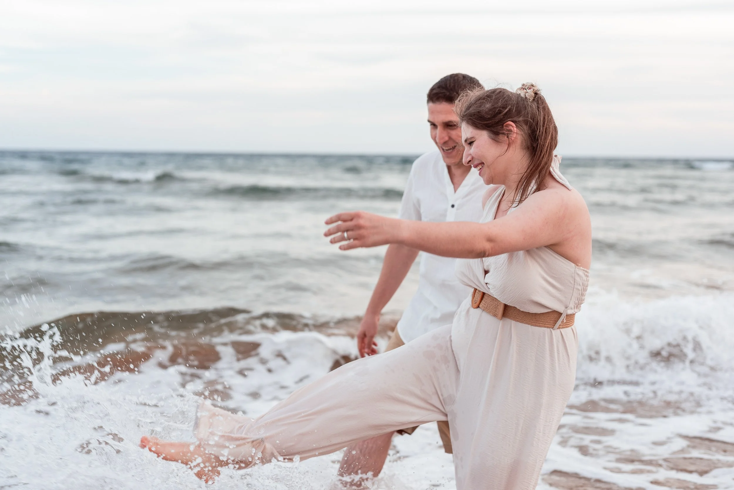 Una pareja sonriente disfrutando en la playa, chapoteando en el agua con olas suaves y un cielo nublado.