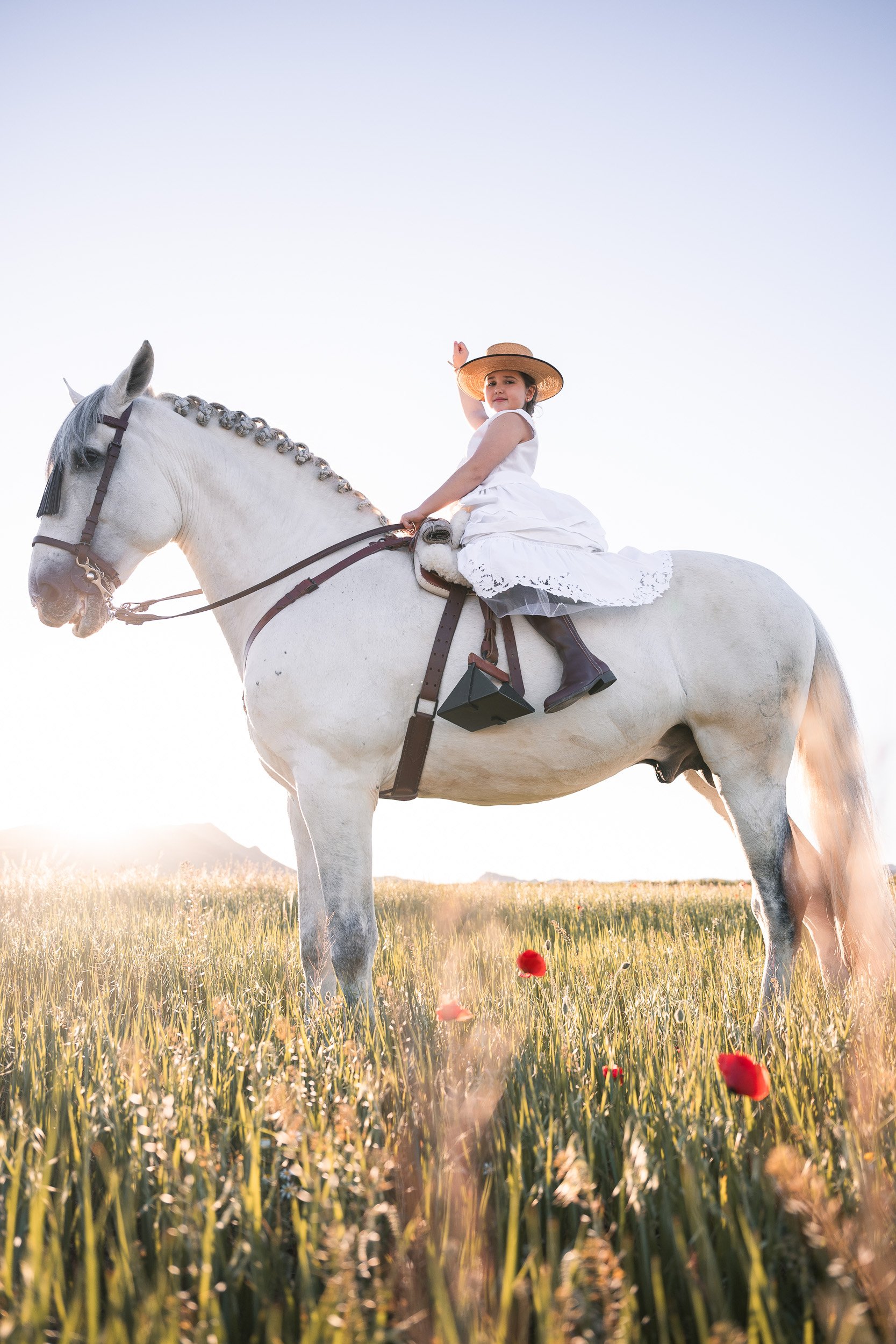 Una niña vestida con un vestido blanco y sombrero sentado en un caballo blanco en un campo con flores rojas, al atardecer.