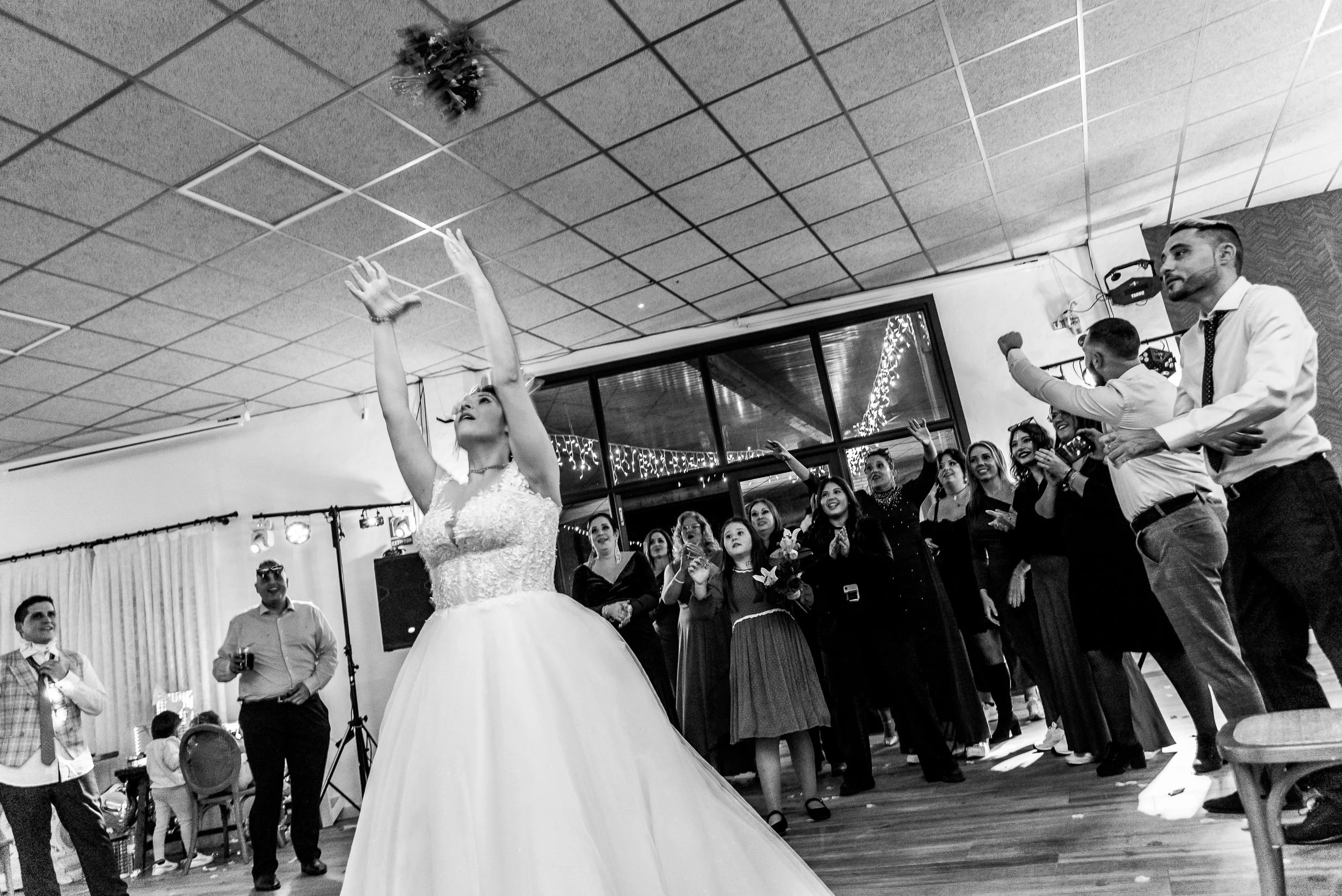 Una novia bailando en una celebración de boda con muchos invitados sonriendo y tomándose fotos en un espacio cerrado decorado con luces y adornos navideños.