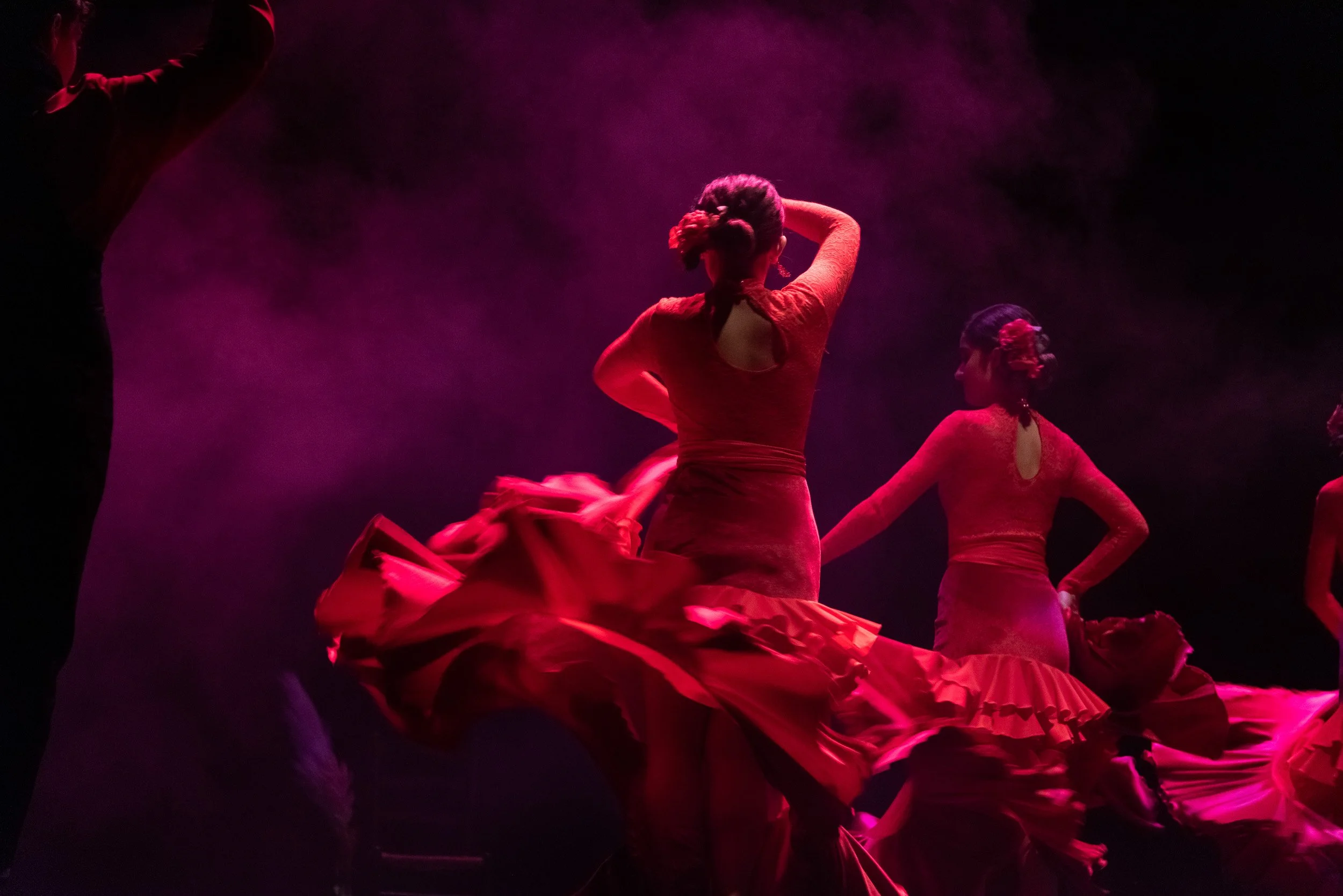 Mujeres bailarinas usando faldas rojas en una presentación de danza en un escenario con iluminación rosa y humo de fondo.