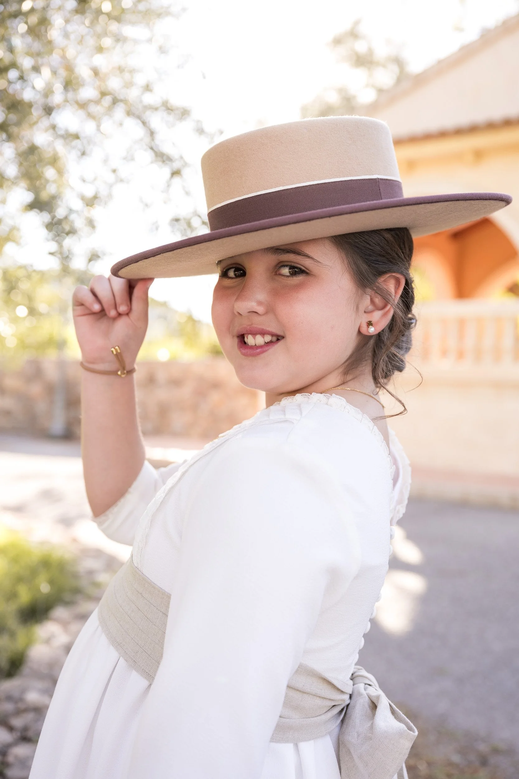 Niña sonriendo y usando sombrero beige y vestido blanco en un espacio al aire libre, con fondo de árboles y casa.