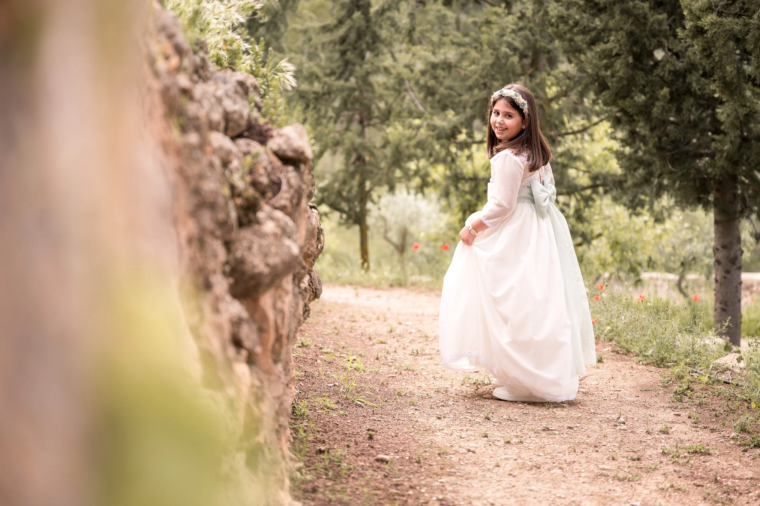Una niña vestida de comunión y diadema de flores en un entorno natural con árboles y flores en el fondo.