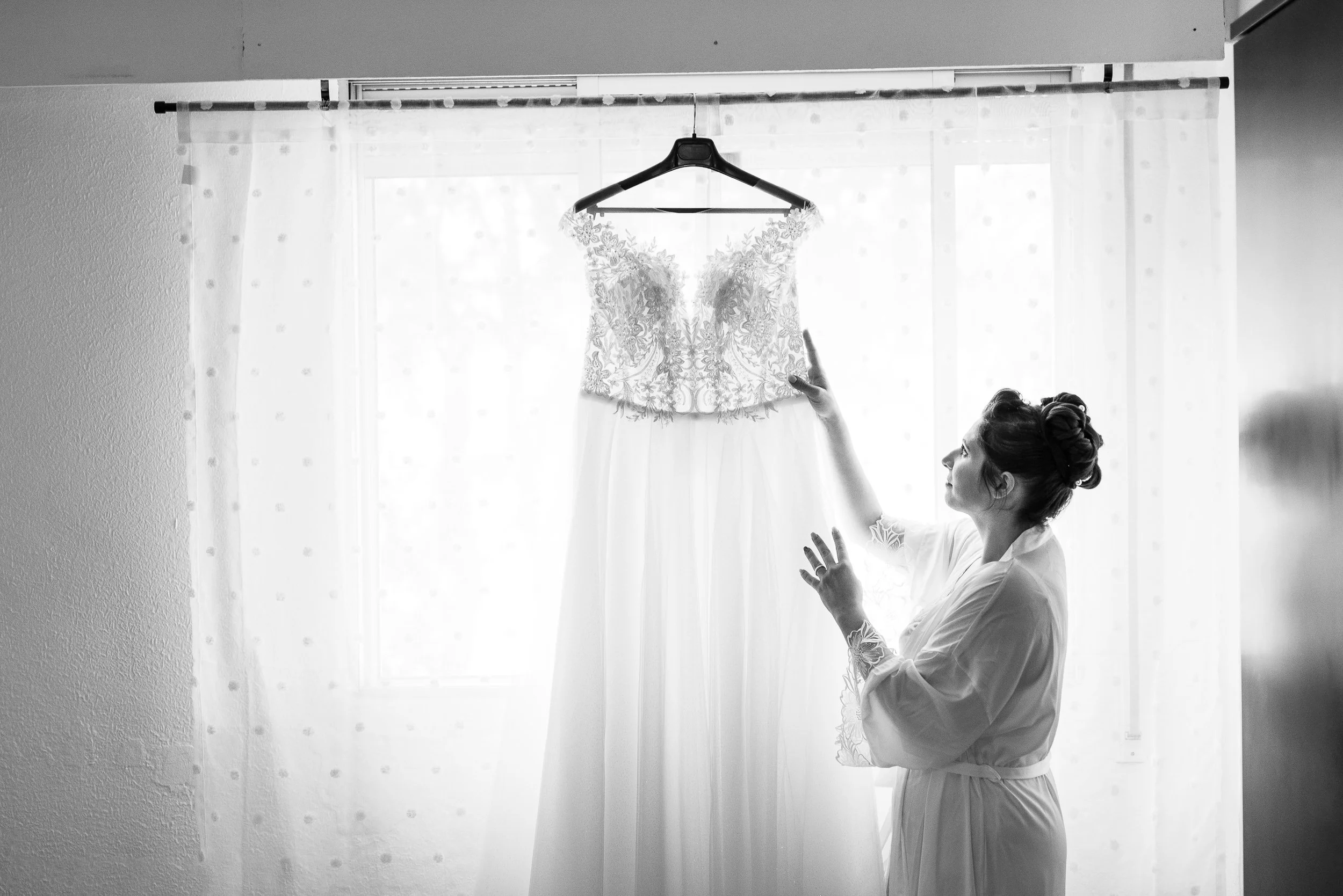 Mujer con peinado recogido, de perfil, con bata, mira una etiqueta en un vestido de novia que cuelga de una percha, frente a una ventana con cortinas blancas.