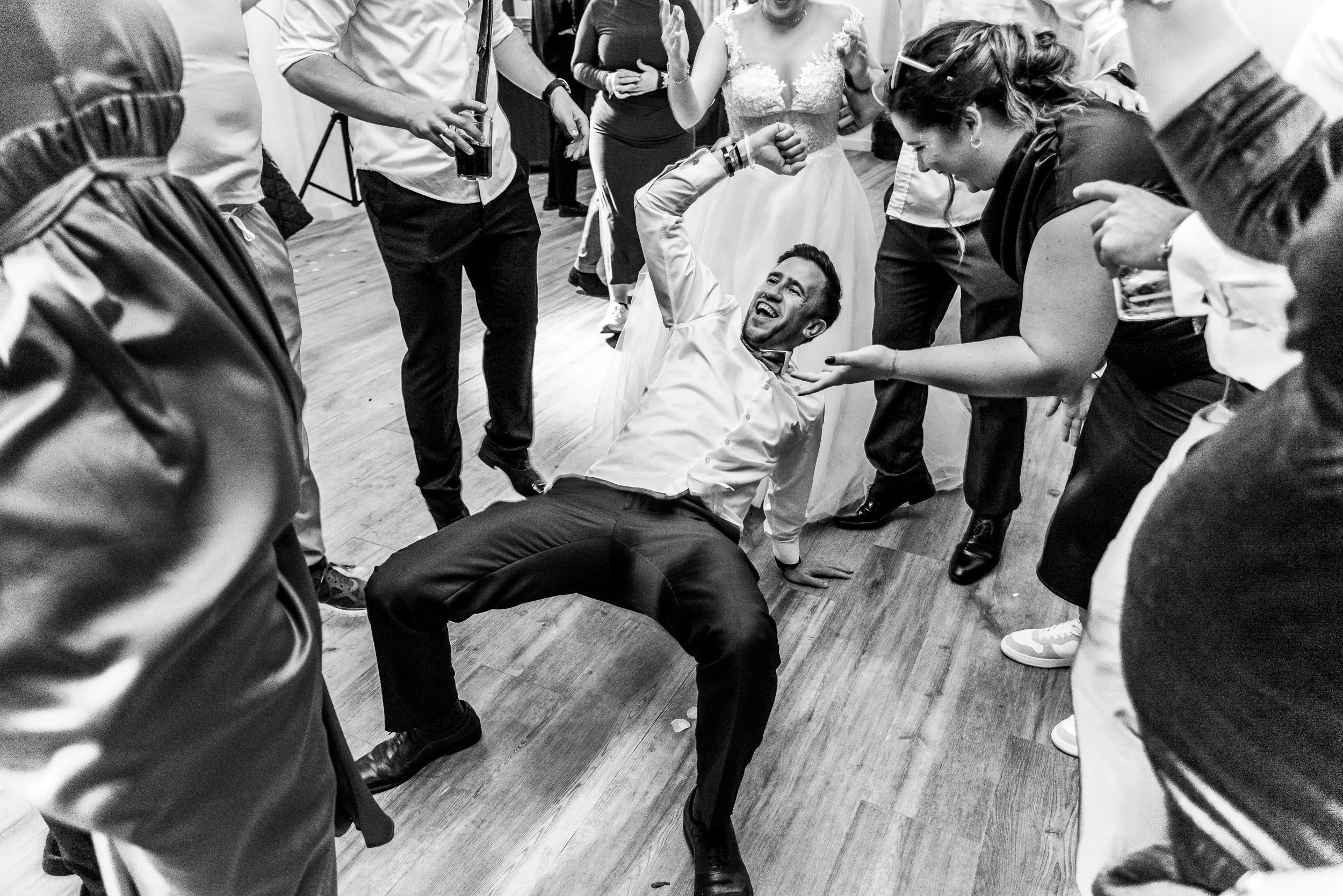 Foto en blanco y negro de una boda, con varias personas bailando y sonriendo. Un hombre está en el suelo en una pose de baile, rodeado de amigos que lo animan y disfrutan del momento.