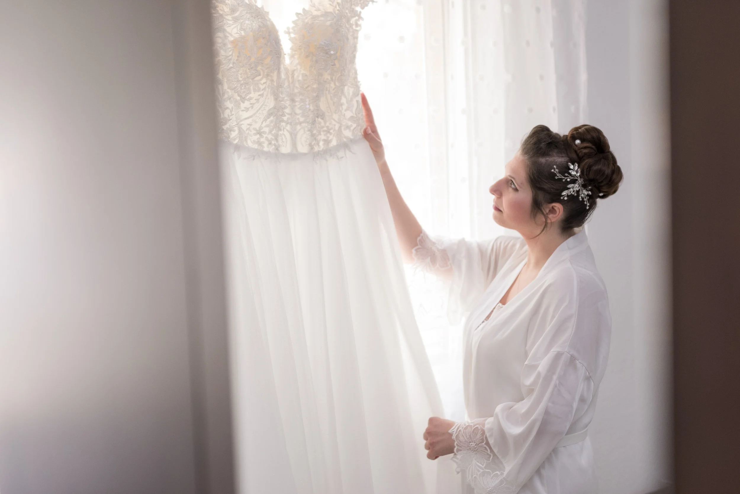 Una mujer con cabello peinado, usando un vestido de novia, mira una prenda de vestido de novia colgada en la ventana.