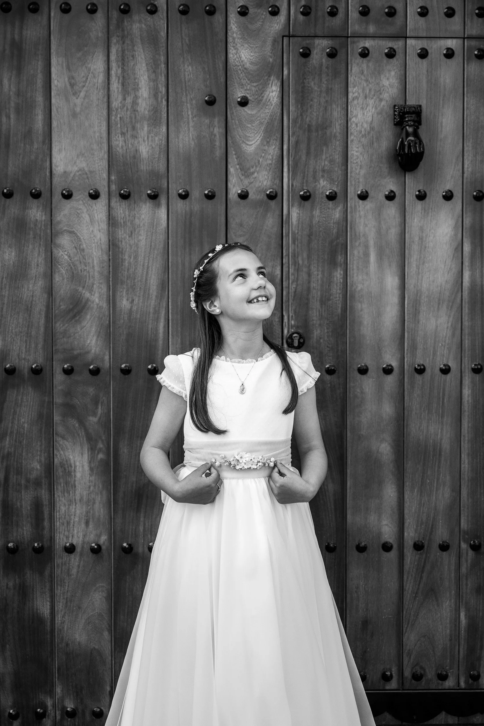 Una niña con vestido de comunión y diadema, sonriendo y tocando su vestido, frente a una puerta de madera con clavos decorativos.