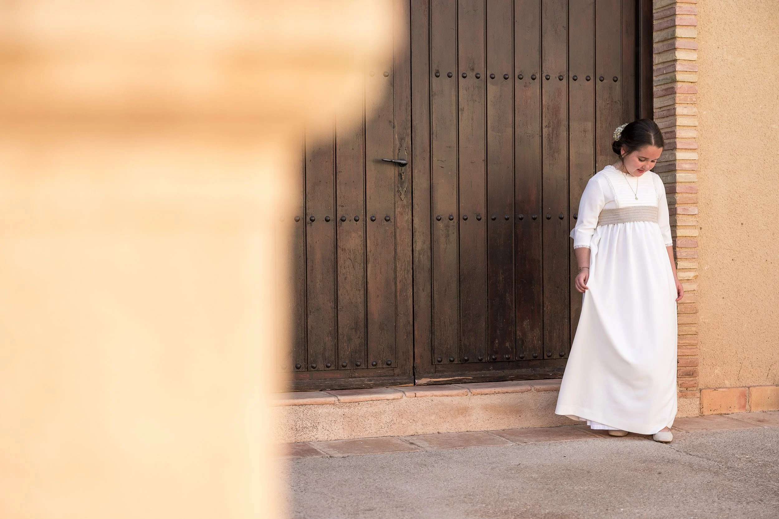 Niña con vestido blanco y accesorios en el cabello, sonriendo y mirando hacia abajo, de pie frente a una puerta de madera con pared de ladrillos y pared de color beige