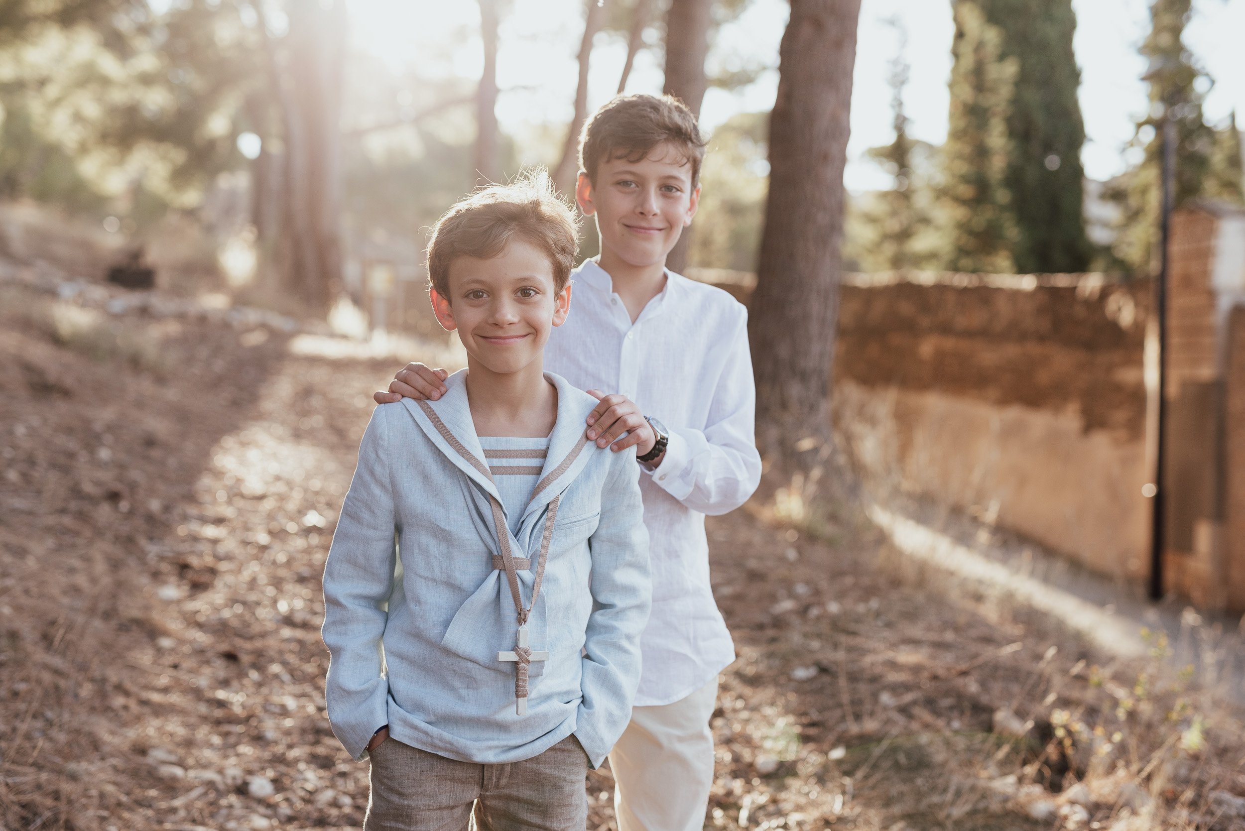Fotografía de dos hermanos de comunión sonriendo y mirando a cámara en un paisaje de árboles y bosque al atardecer.