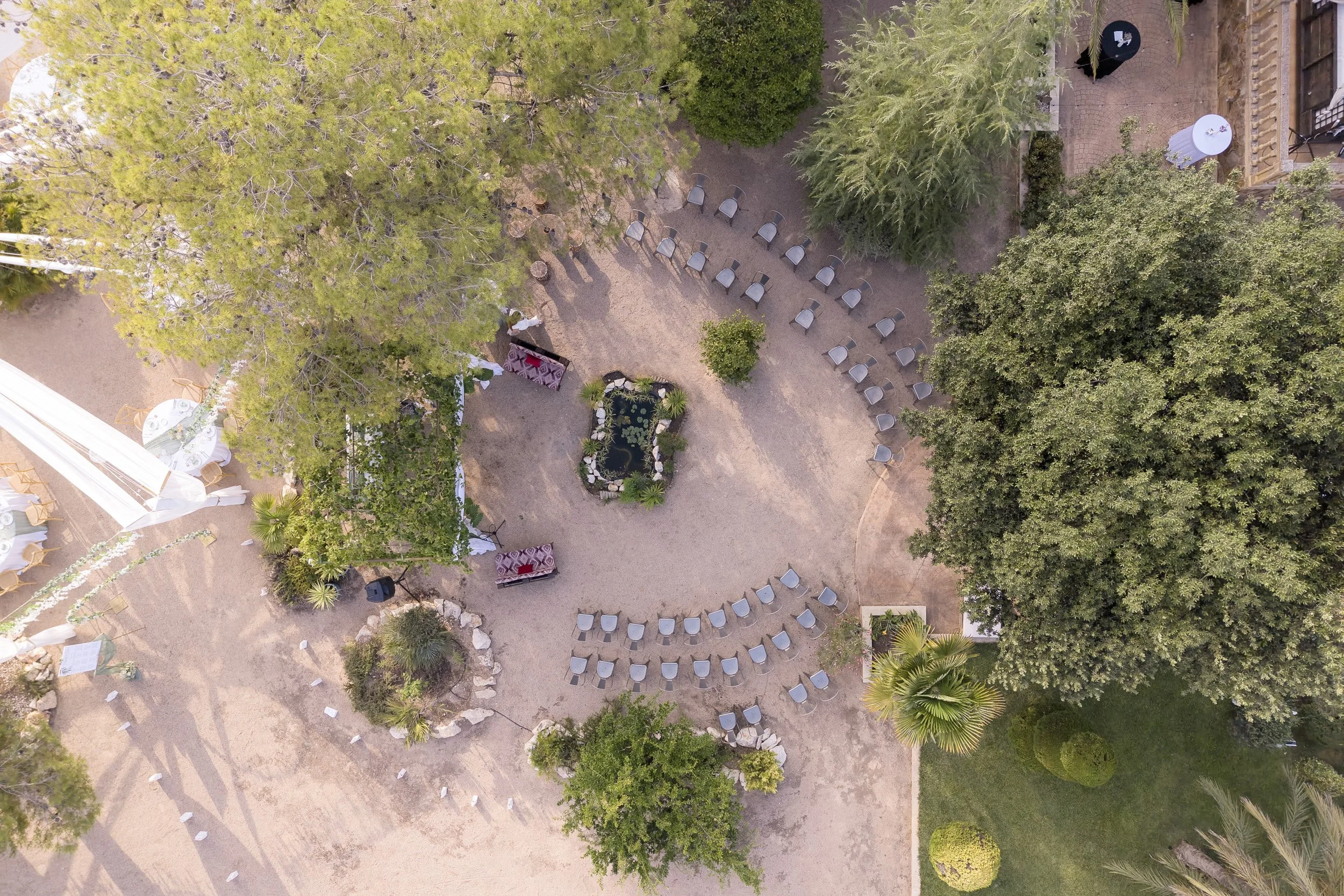 Vista aérea de una boda en un jardín con árboles y sillas dispuestas en círculo y en fila, además de algunas décoraciones y un área con plantas acuáticas en el centro.