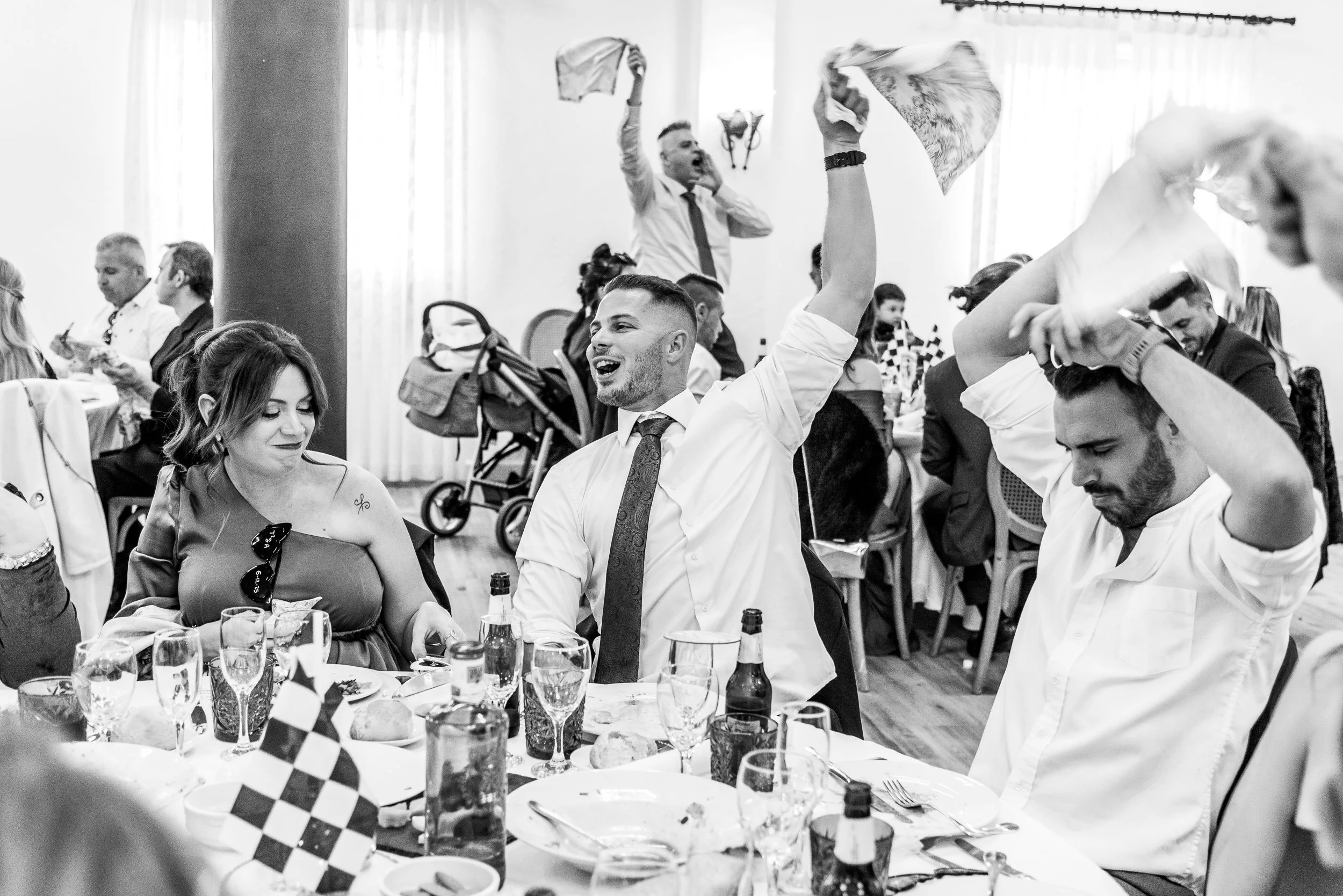 Personas en una celebración, levantando pañuelos y sonriendo en un ambiente festivo, con mesas y bebidas.