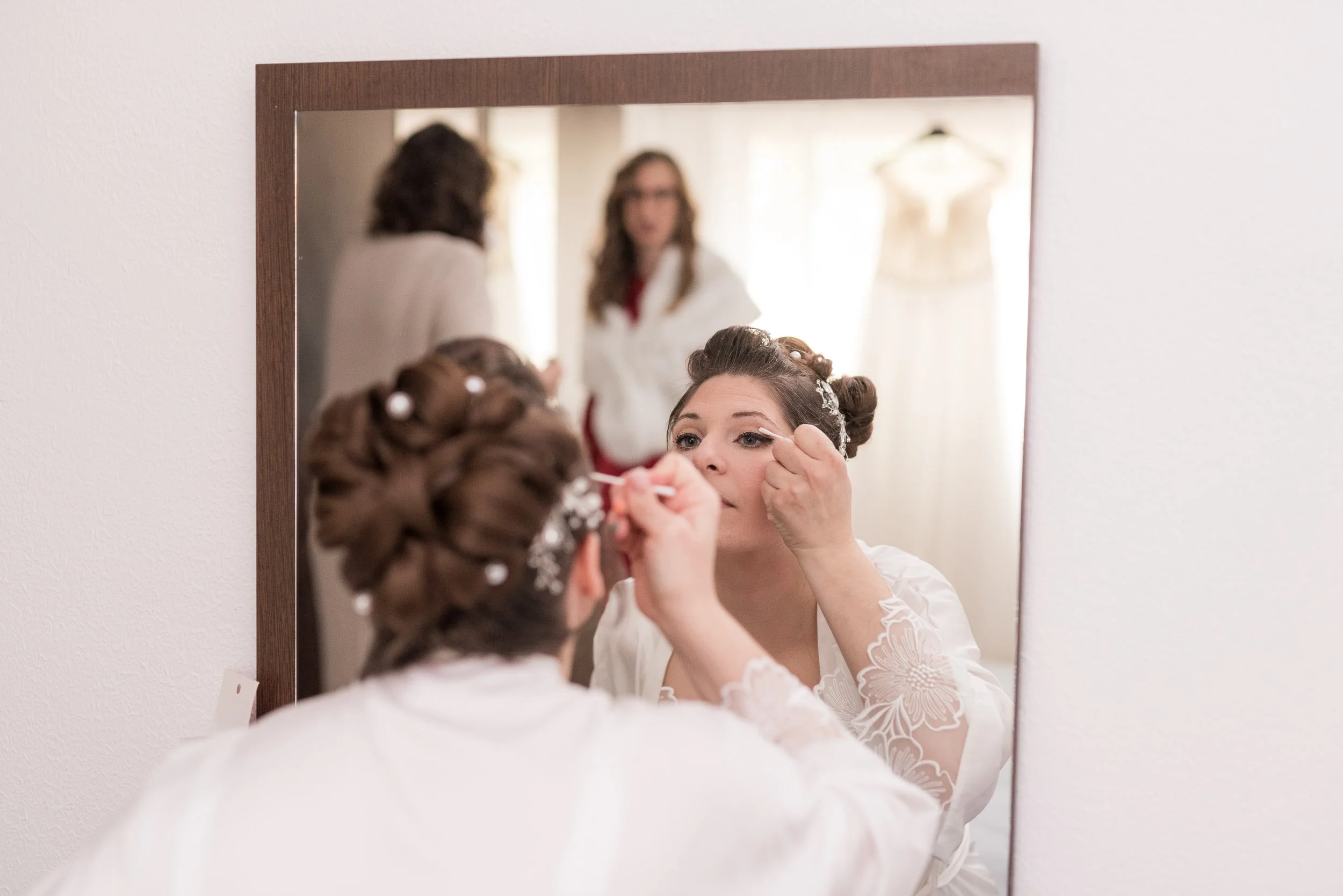 Mujer peinándose frente a un espejo, con cabello rizado adornado con accesorios y vestida con un vestido blanco con detalles de encaje, en un entorno con luz natural.