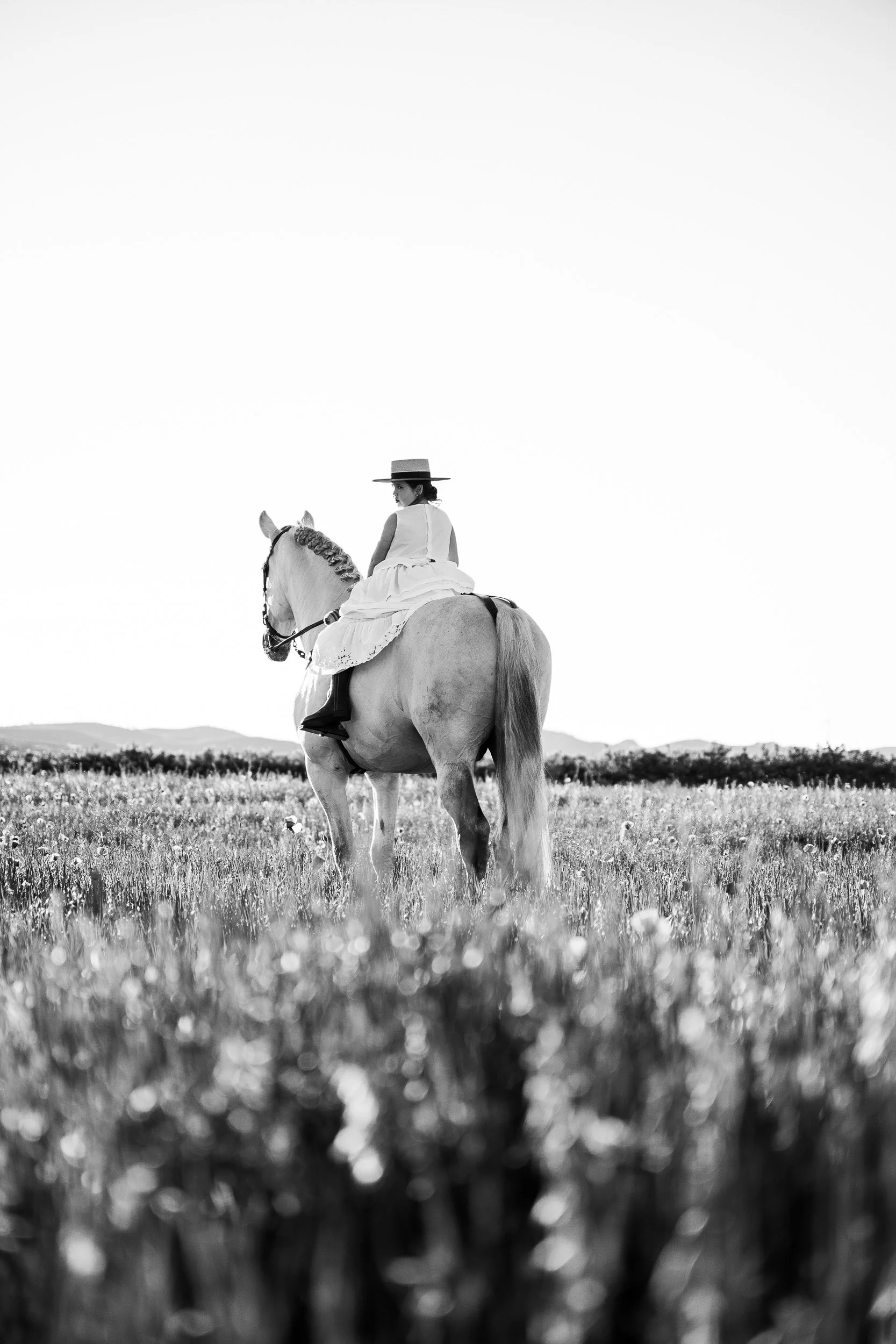 Mujer en vestido blanco y sombrero de ala ancha montando un caballo en un campo con fondo de montañas.