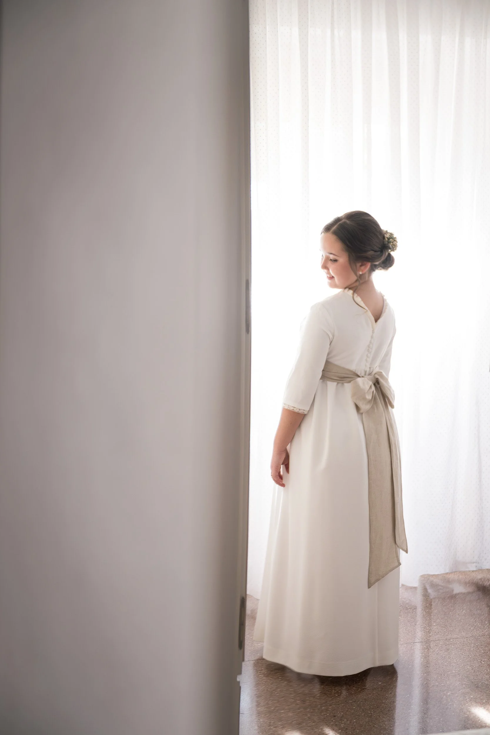Una joven vestida de comunión, en una habitación con cortinas blancas y luz natural.
