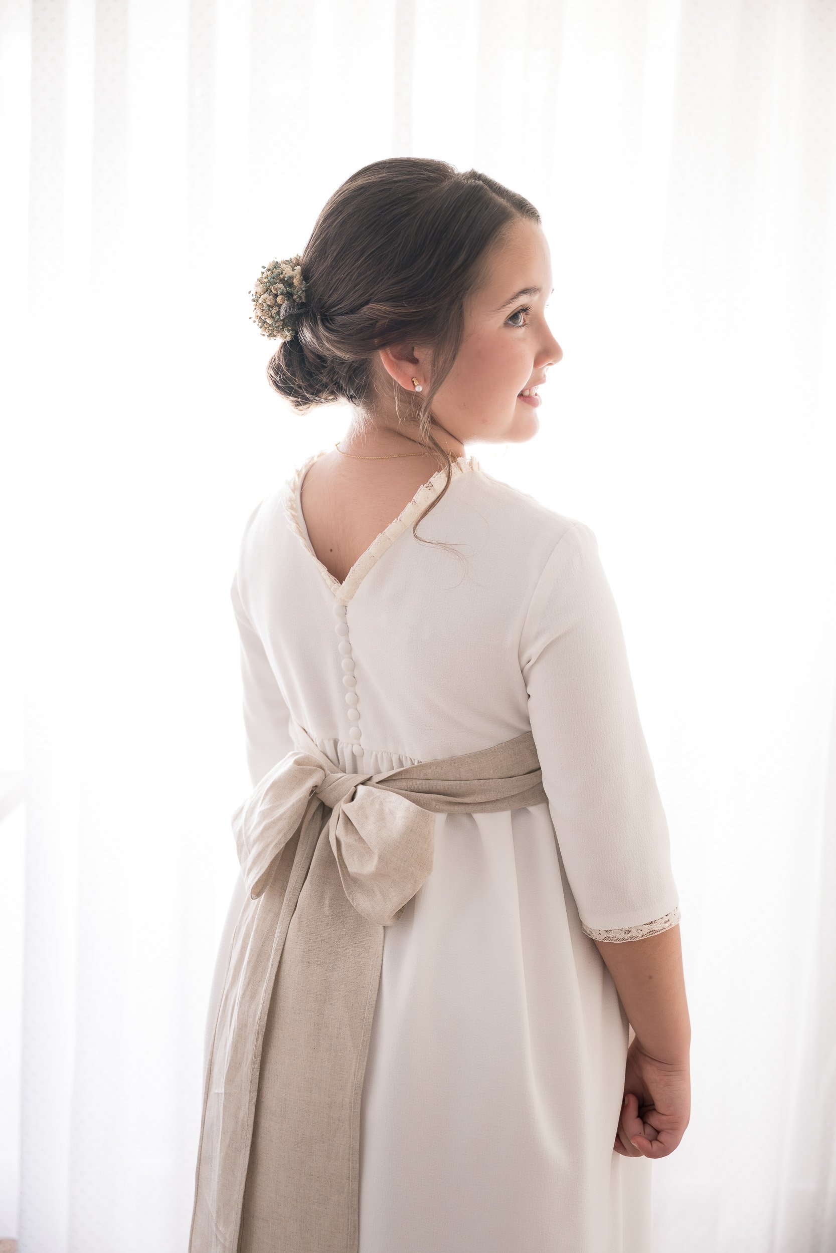 Mujer joven vista desde un lado, con cabello peinado en bollo adornado con flores pequeñas, vestida con blusa blanca y cinturón beige, en un fondo de cortinas blancas.