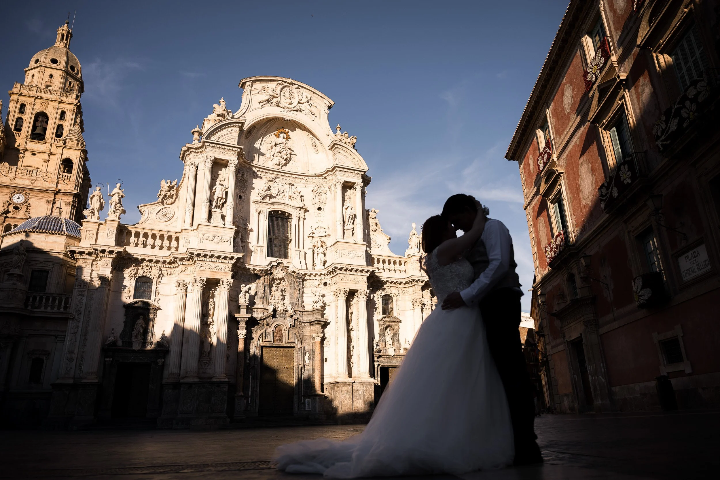 Pareja de novios besándose frente a la catedral de Murcia en un día soleado, en sombra y alto contraste de luces.