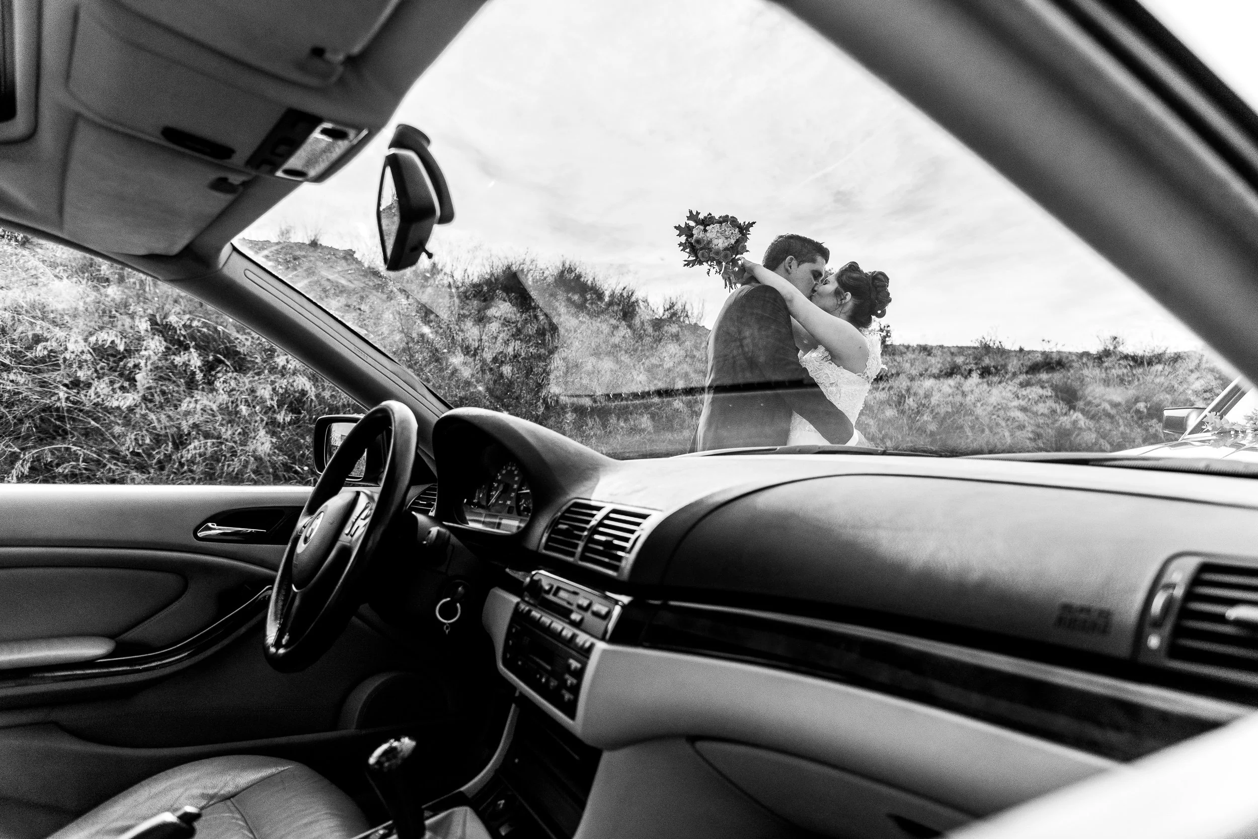 La foto muestra la vista desde el interior de un automóvil, mirando hacia afuera a través del parabrisas. Se observa a una pareja de novios, un hombre y una mujer, compartiendo un beso en un entorno natural, mientras el coche está detenido en un área