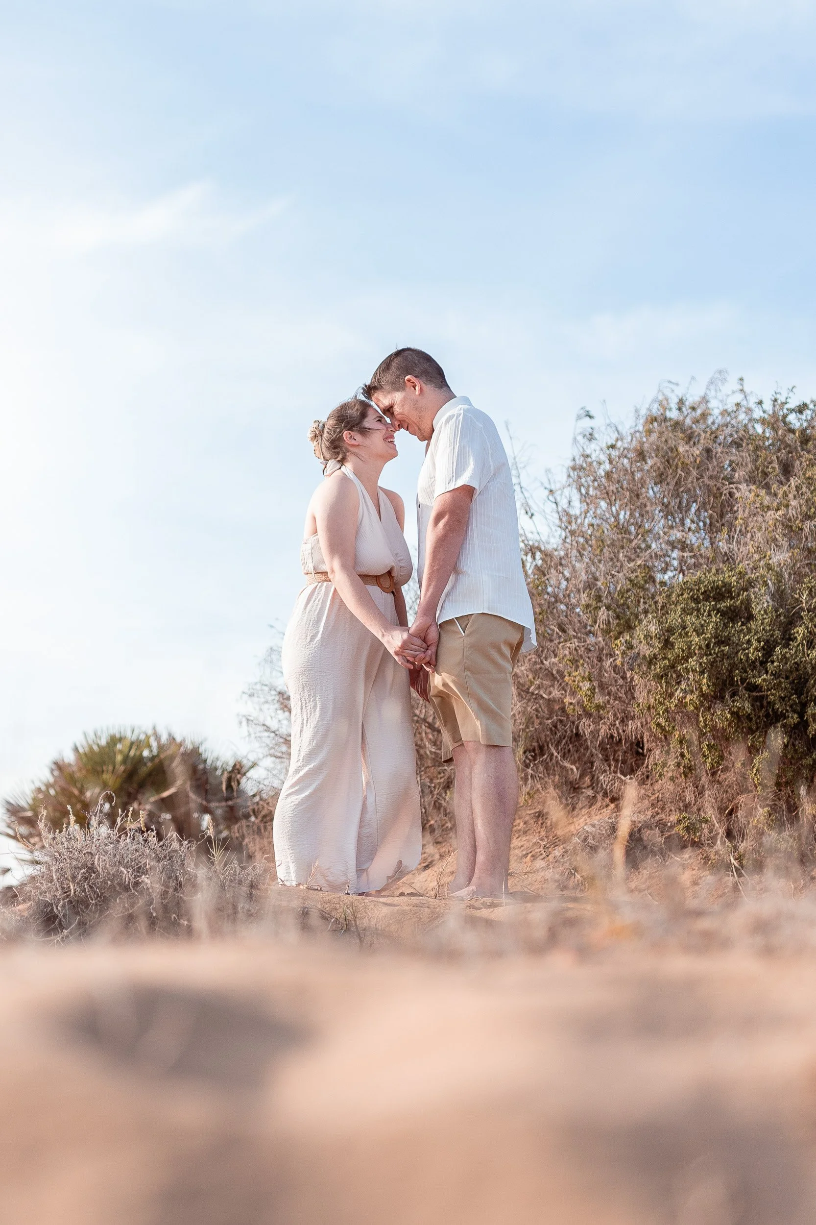Pareja joven en un paisaje de playa natural, sosteniéndose de las manos y tocándose las frentes con una expresión de felicidad.