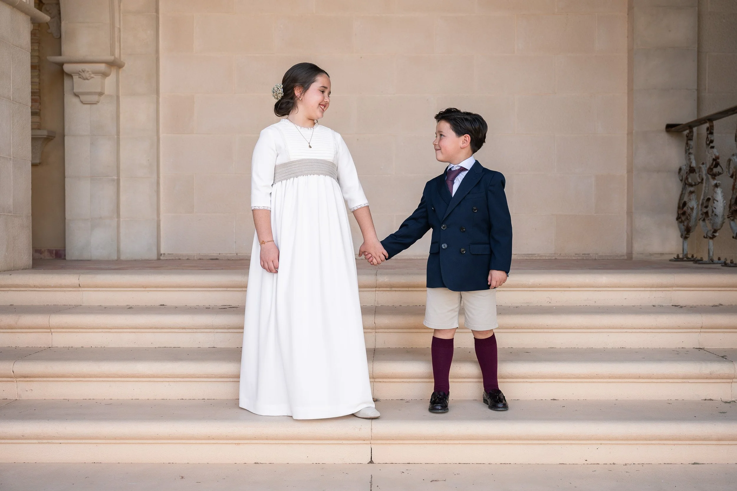 Una niña vestida con vestido largo y un niño con traje formal y pantalones cortos, tomados de la mano y sonriendo en las escaleras de un edificio de piedra.