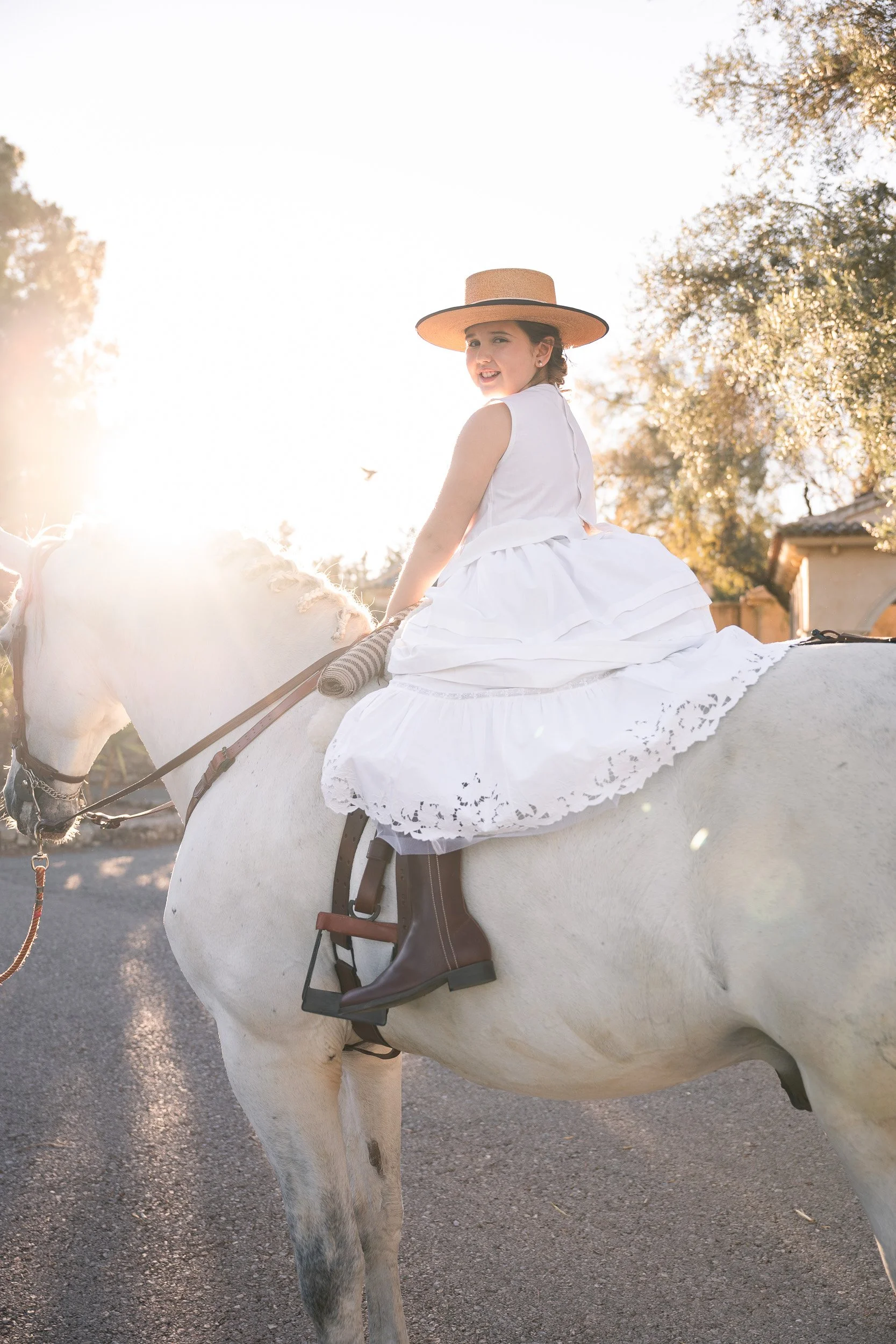 Joven montando a caballo, viste vestido blanco y sombrero de paja, en un entorno al atardecer con árboles y casas en el fondo.