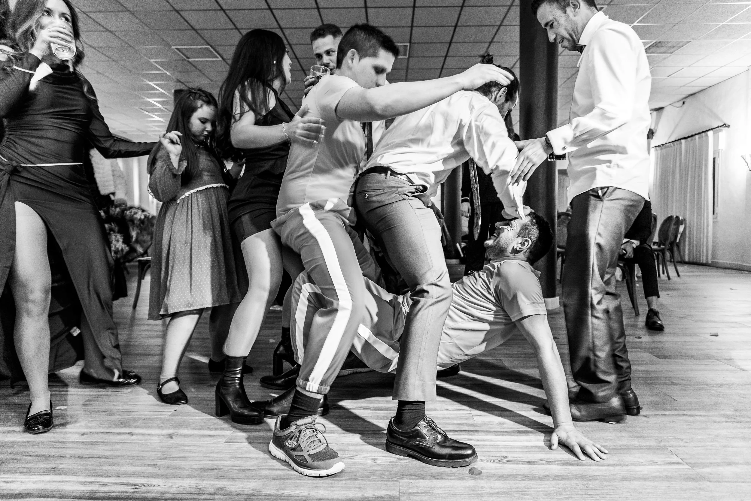 Grupo de personas participando en un juego de coordinación y equilibrio, una persona en el suelo con una mano en el piso mientras otra persona le ayuda a levantarse, en un espacio con piso de madera y techo de plafón.