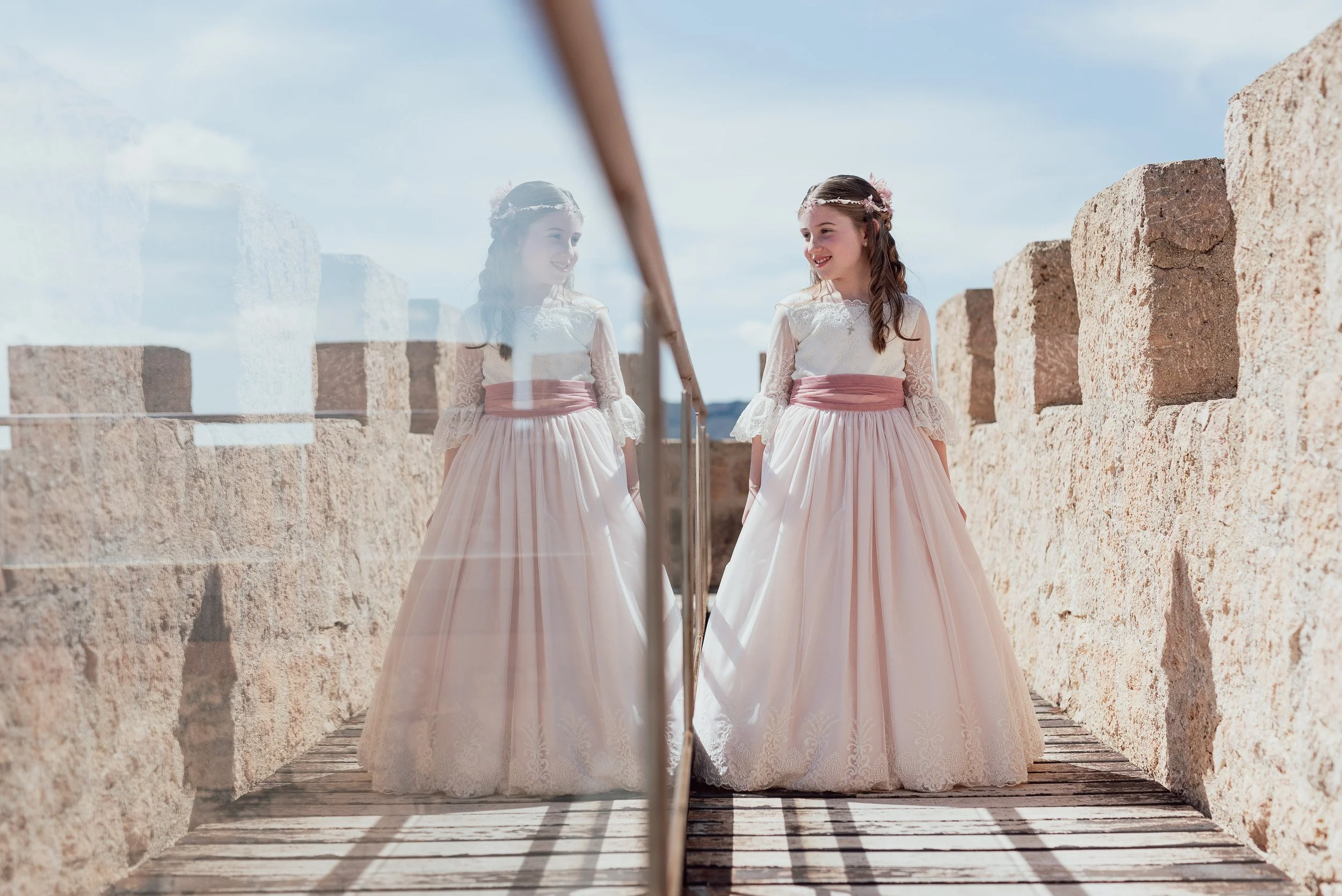 Una niña vestida de comunión paseando por un castillo mientras que es reflejada por el cristal de una pasarela del castillo.